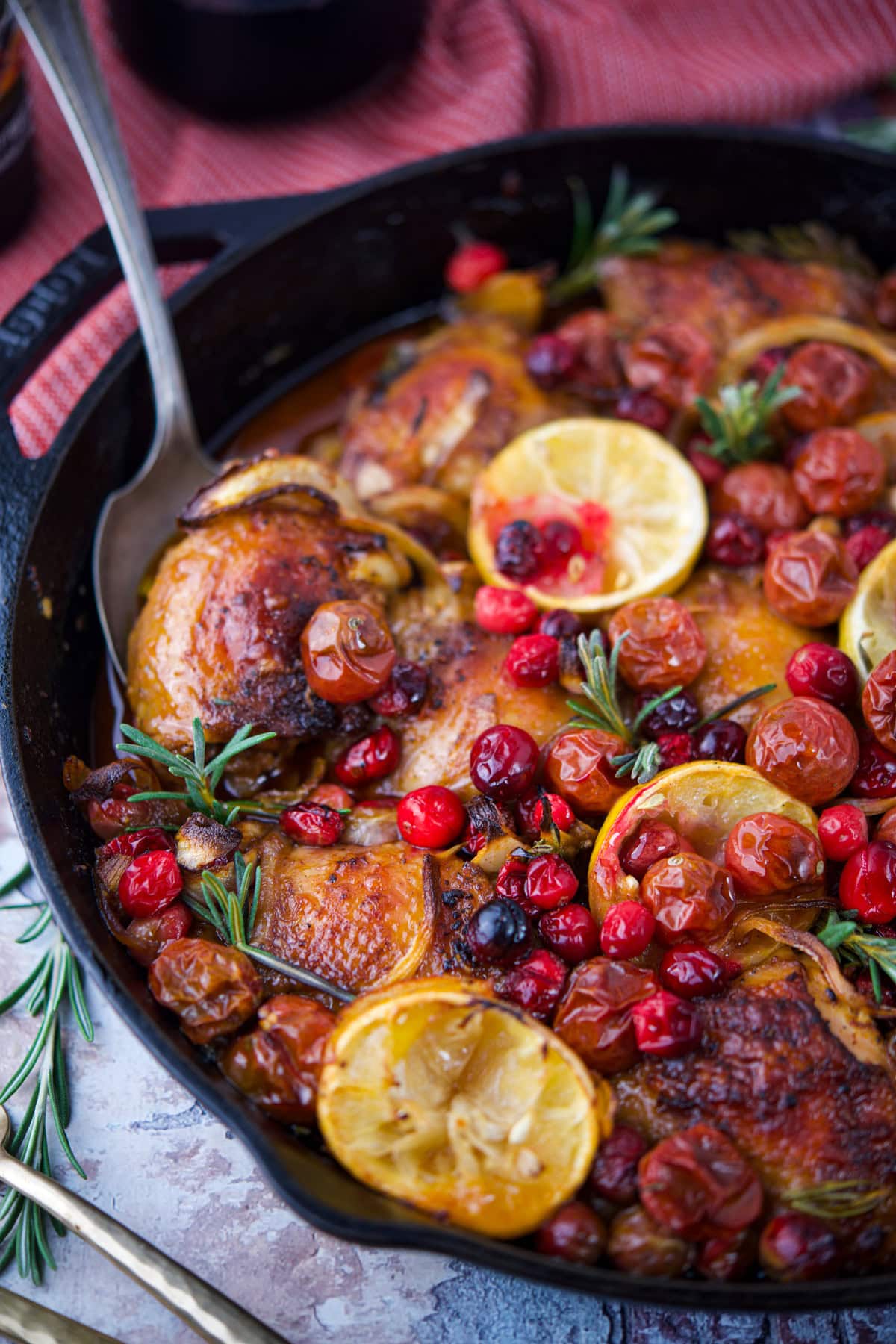 A skillet filled with roasted Cranberry Lemon Chicken, garnished with lemon slices, cherry tomatoes, red cranberries, and fresh rosemary sprigs. A serving spoon rests inside the pan. The dish looks vibrant and appetizing.