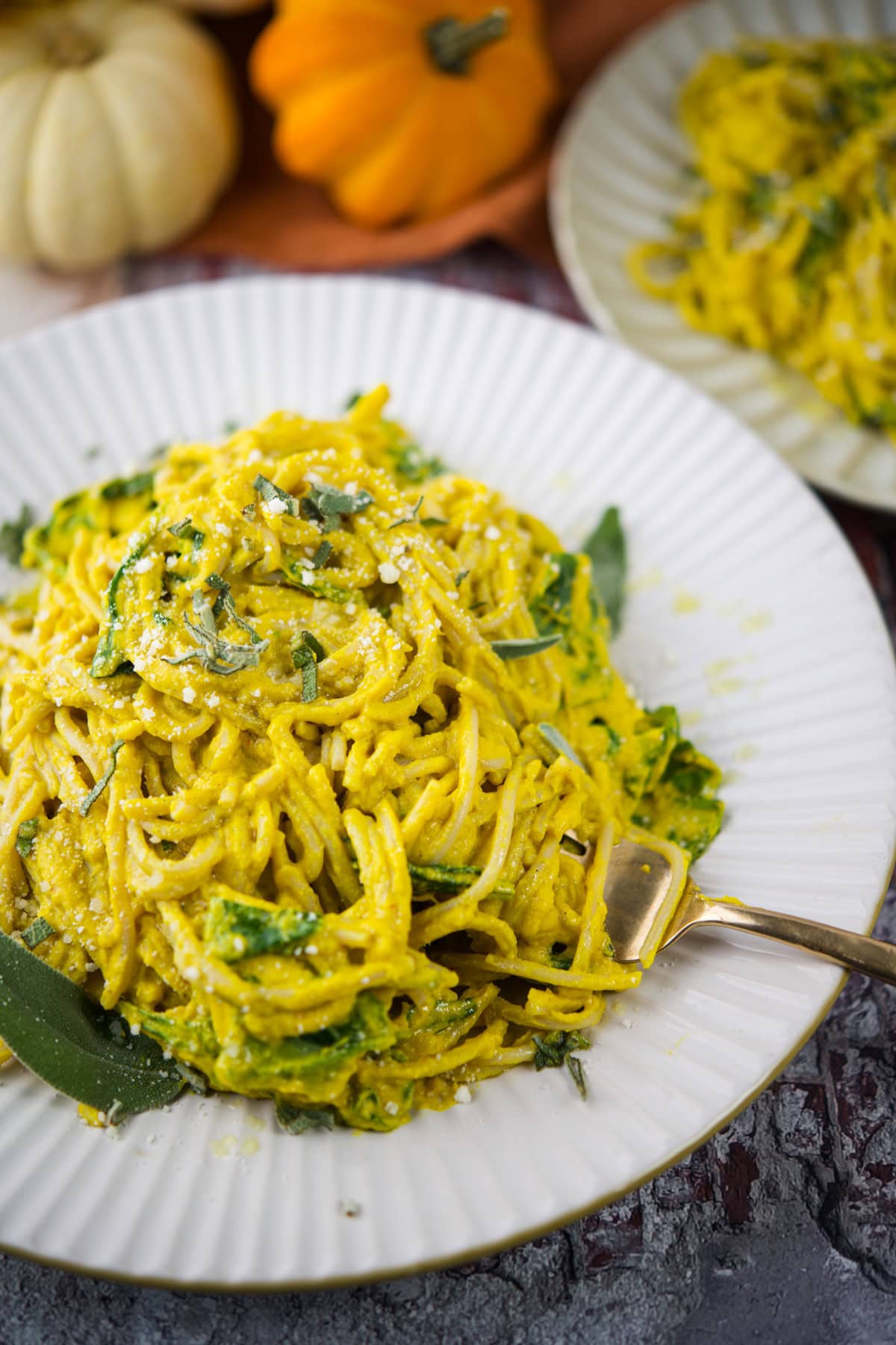 A plate of spaghetti coated in a creamy yellow sauce, garnished with fresh herbs and grated cheese, sits on a white plate with a fork. Pumpkins and another plate of pasta are blurred in the background.