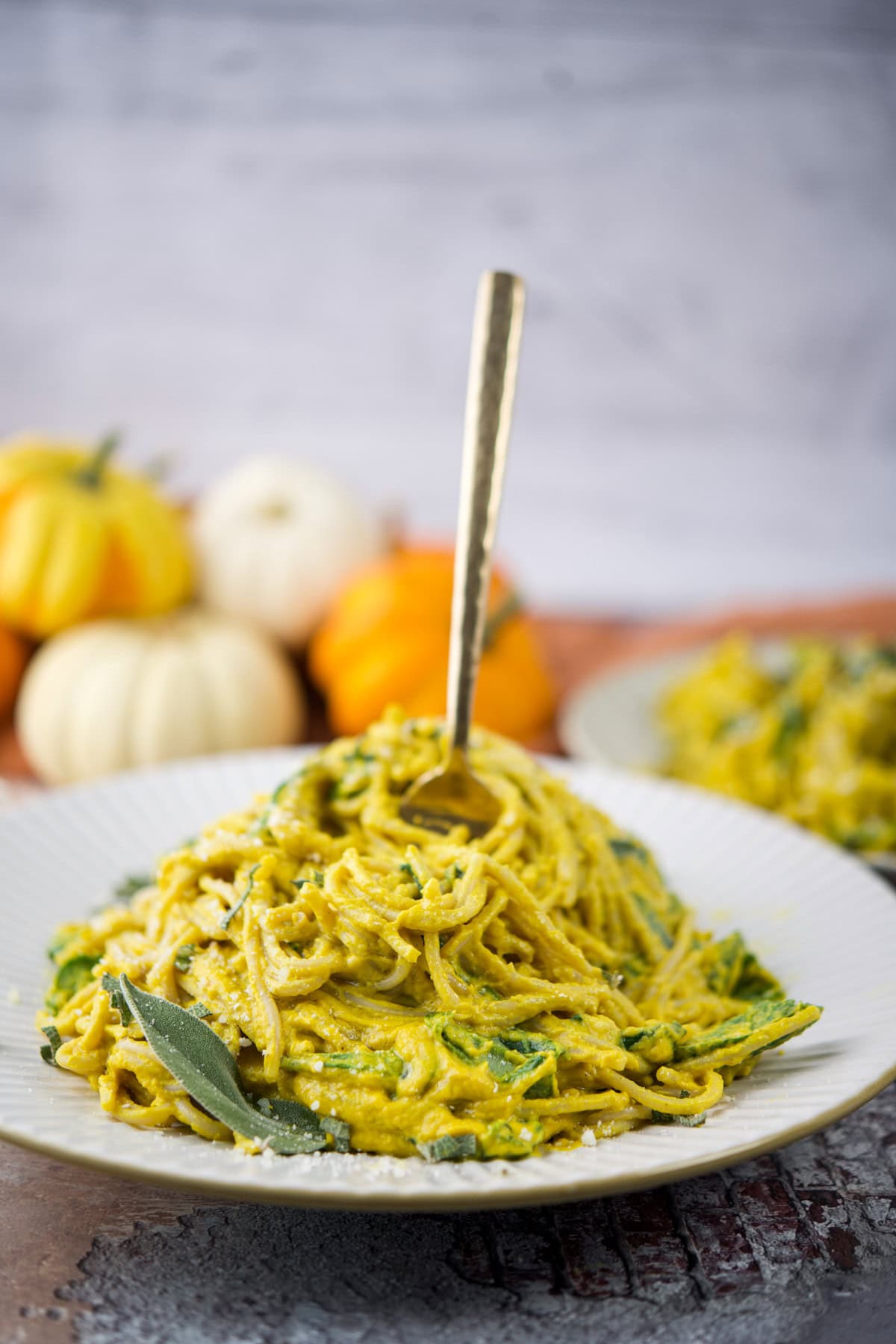 A plate of creamy pasta with green herbs is garnished with a sage leaf and served with a fork inserted in the center. In the blurred background, small decorative pumpkins are visible.
