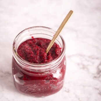 An open glass jar filled with Polish Beet and Horseradish (Ćwikła z Chrzanem) sits on a white marble surface, with a small gold spoon resting inside.