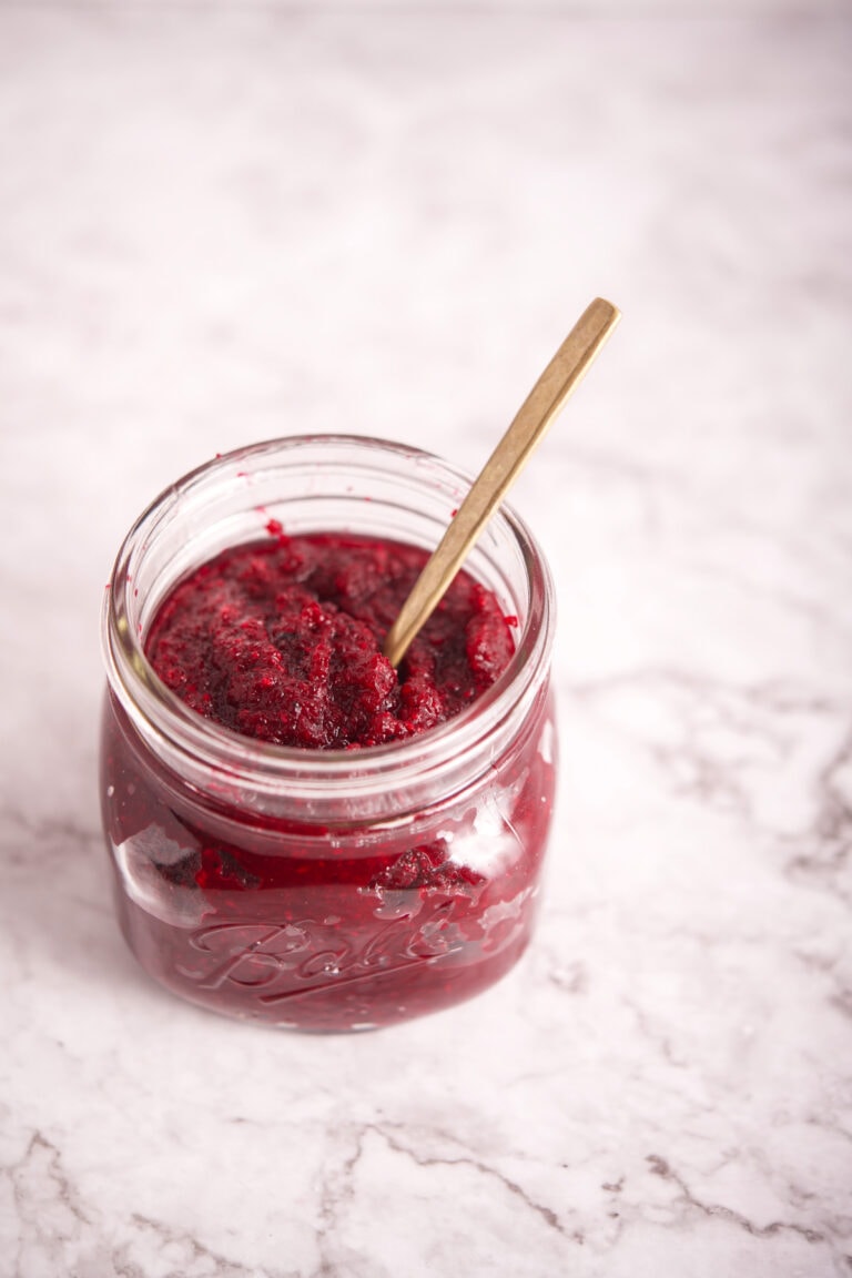 An open glass jar filled with Polish Beet and Horseradish (Ćwikła z Chrzanem) sits on a white marble surface, with a small gold spoon resting inside.