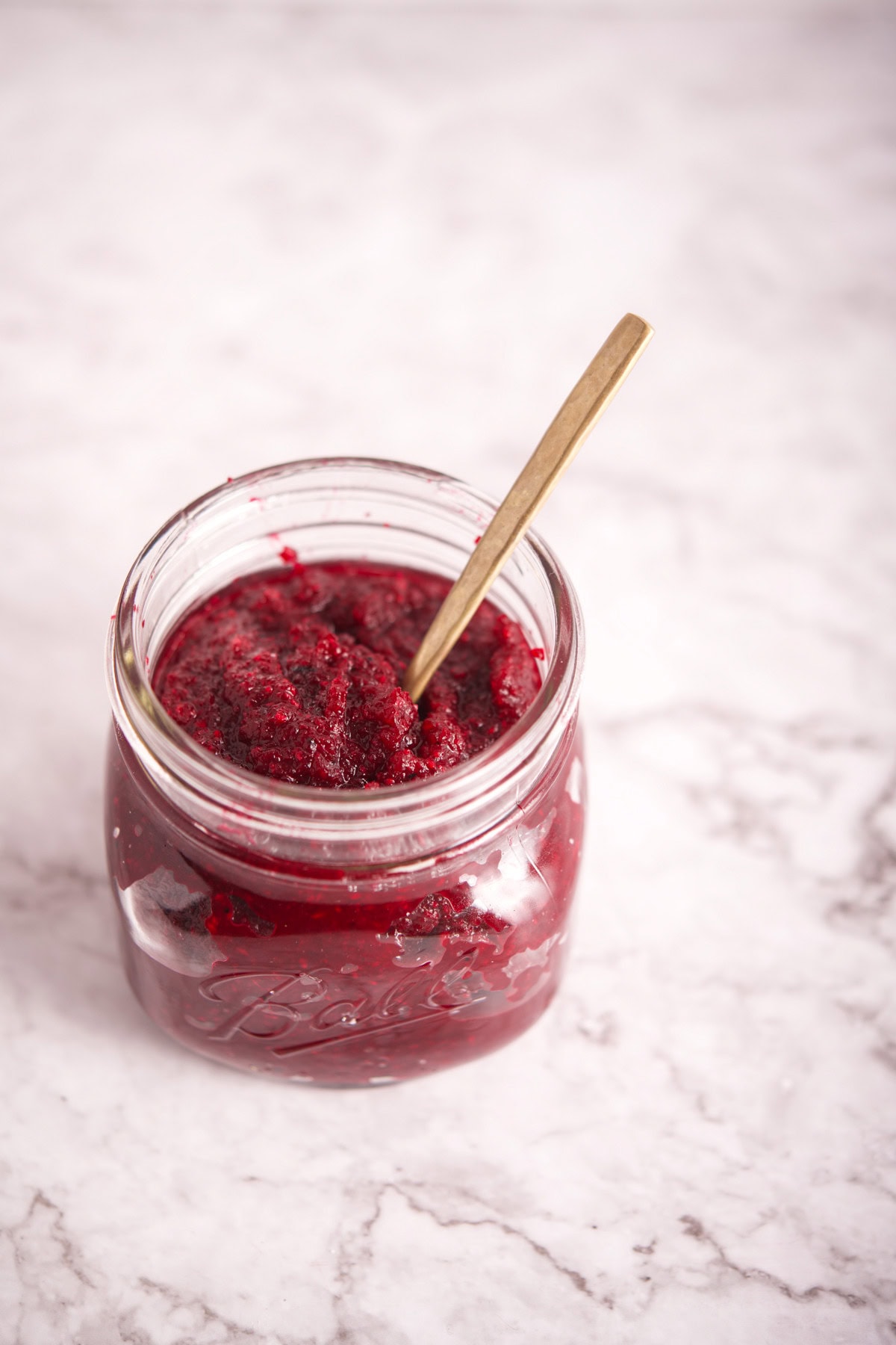 An open glass jar filled with Polish Beet and Horseradish (Ćwikła z Chrzanem) sits on a white marble surface, with a small gold spoon resting inside.