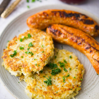 A plate with two Fuczki Polish Sauerkraut Pancakes (Gluten-Free) garnished with parsley, served alongside two grilled sausages. A jar of red sauce and cutlery are visible in the background.