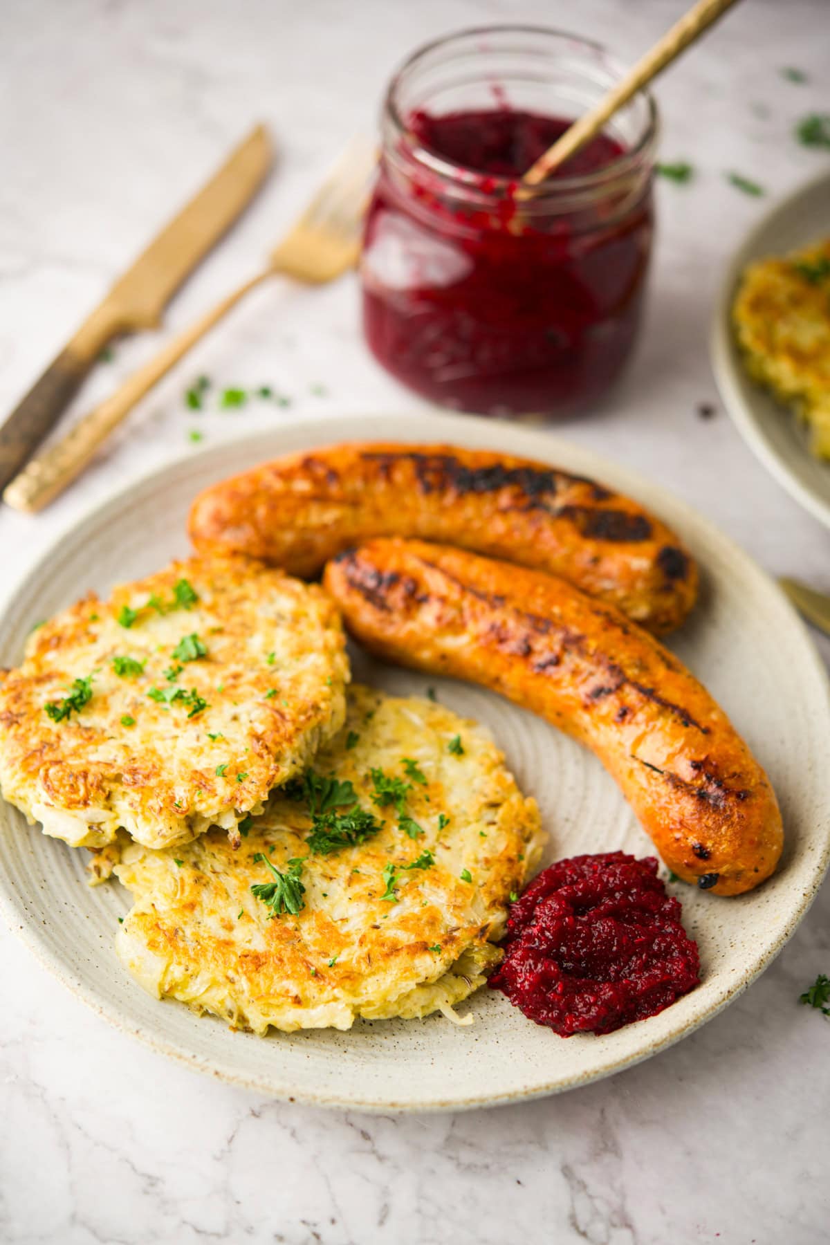 A plate with two browned sausages, two golden potato pancakes topped with herbs, and a dollop of Polish Beet and Horseradish (Ćwikła z Chrzanem). In the background, there&rsquo;s a jar of red sauce with a spoon and some cutlery.