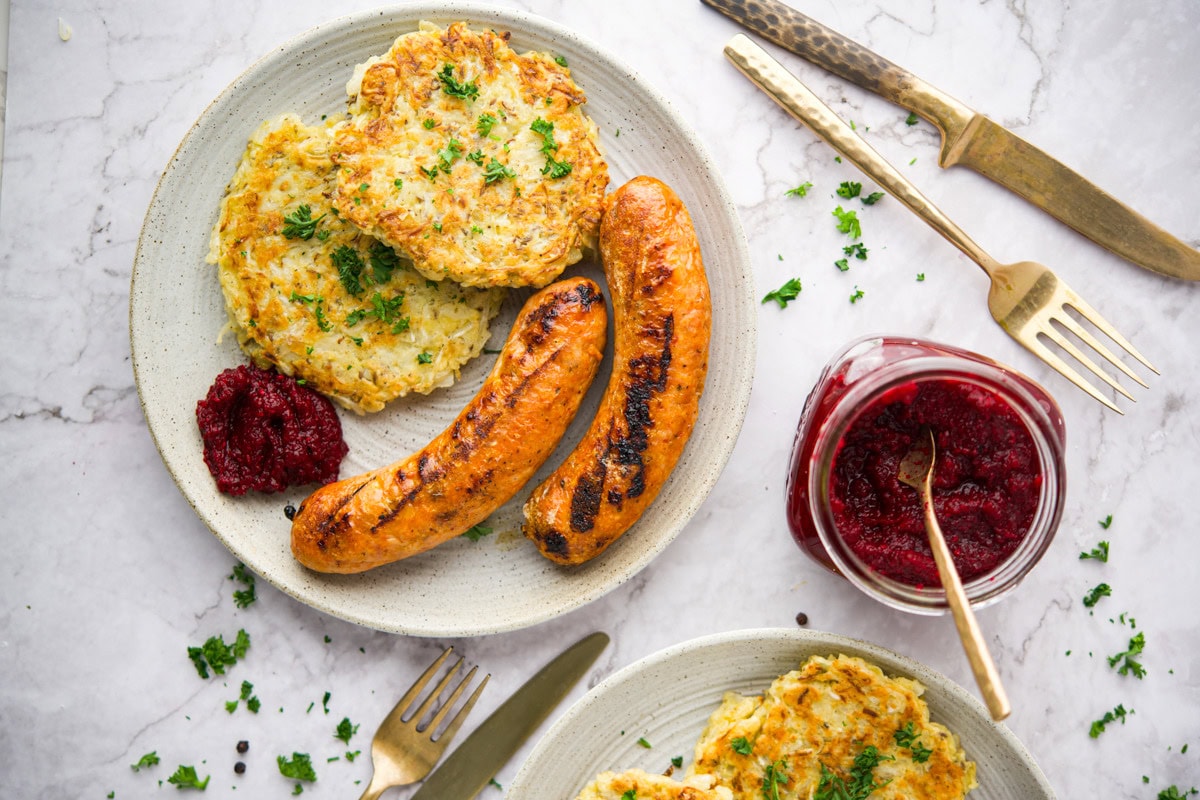 A plate with two grilled sausages, two potato pancakes, a serving of Polish Beet and Horseradish (Ćwikła z Chrzanem), and chopped herbs. Next to the plate are a fork, knife, and a jar of red sauce with a spoon, all on a marble surface.