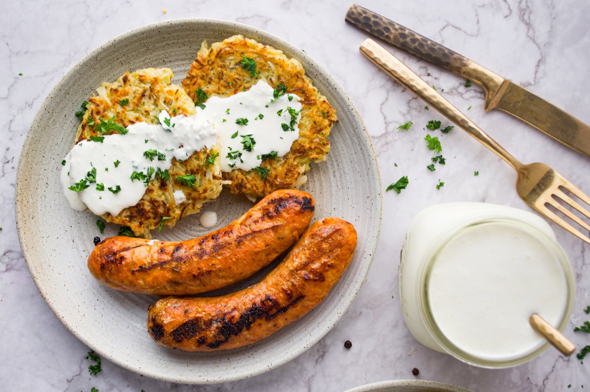 A plate with two grilled sausages and two vegetable fritters topped with white sauce and chopped herbs, next to a glass of milk and gold-colored cutlery on a marble surface.