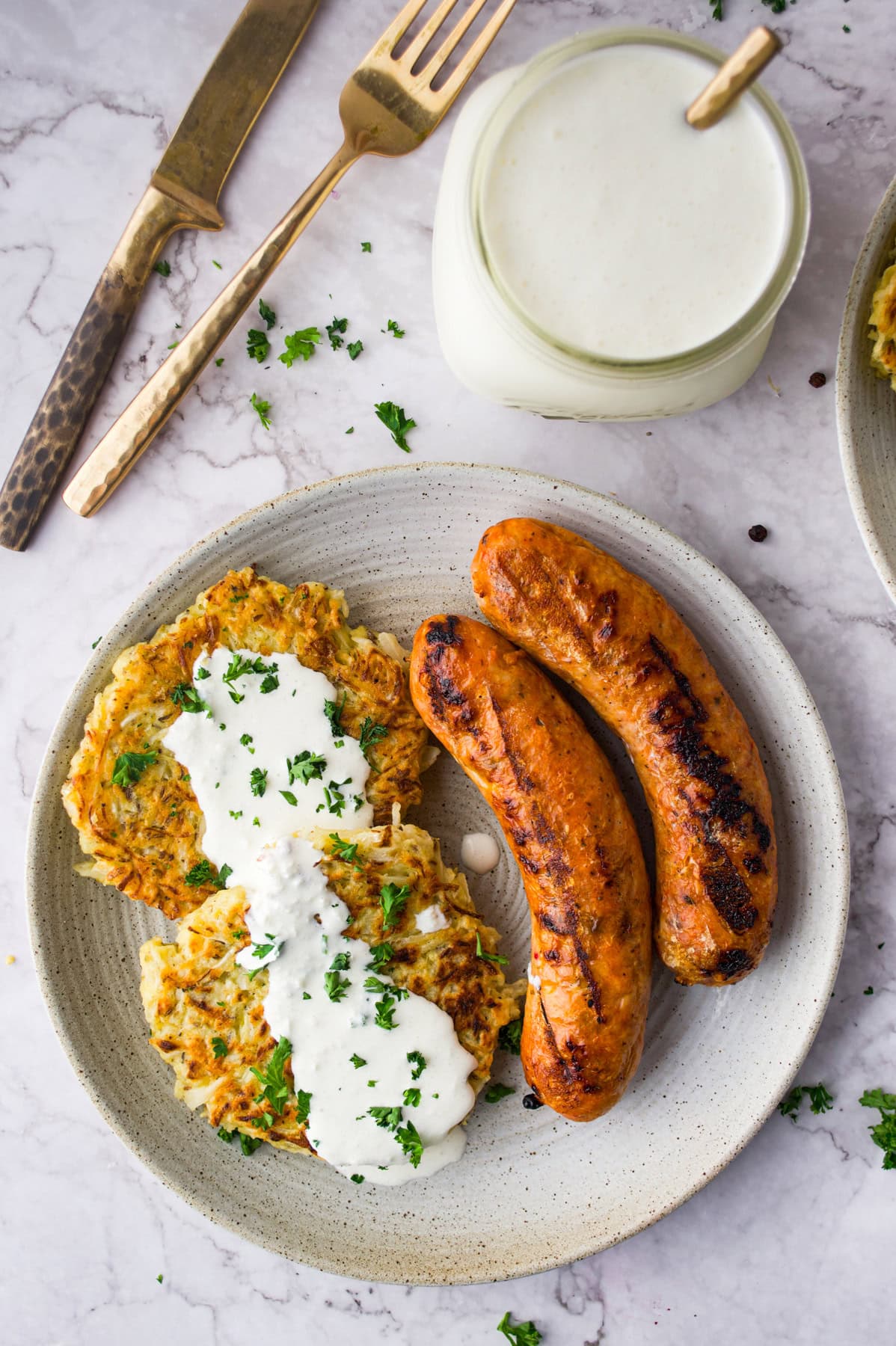 A plate with two grilled sausages and two potato pancakes topped with white sauce and chopped herbs. A jar of sauce with a spoon and a fork and knife are placed nearby on a marble surface.