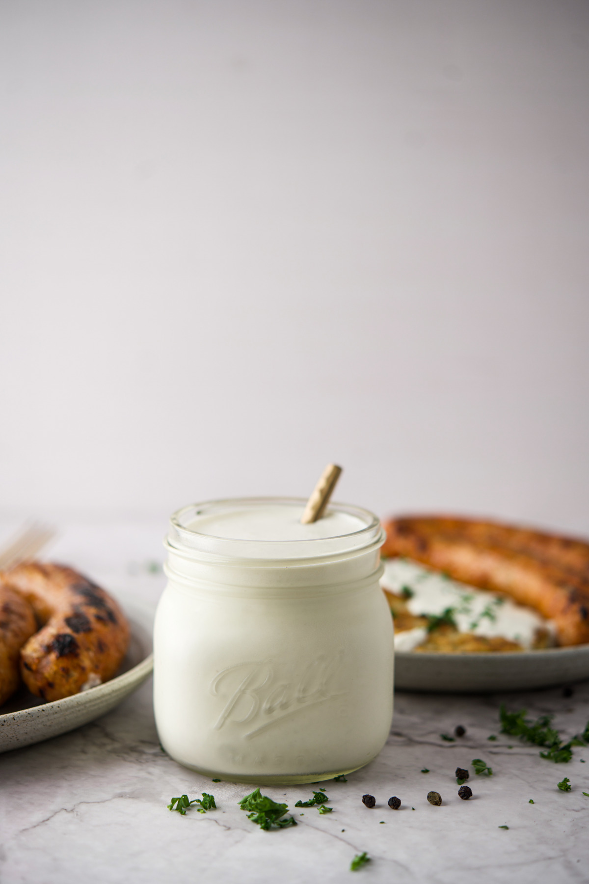 A glass jar filled with Polish Homemade Horseradish Condiment (Chrzan) and a spoon, placed in front of plates with grilled sausages and garnished herbs on a light surface.