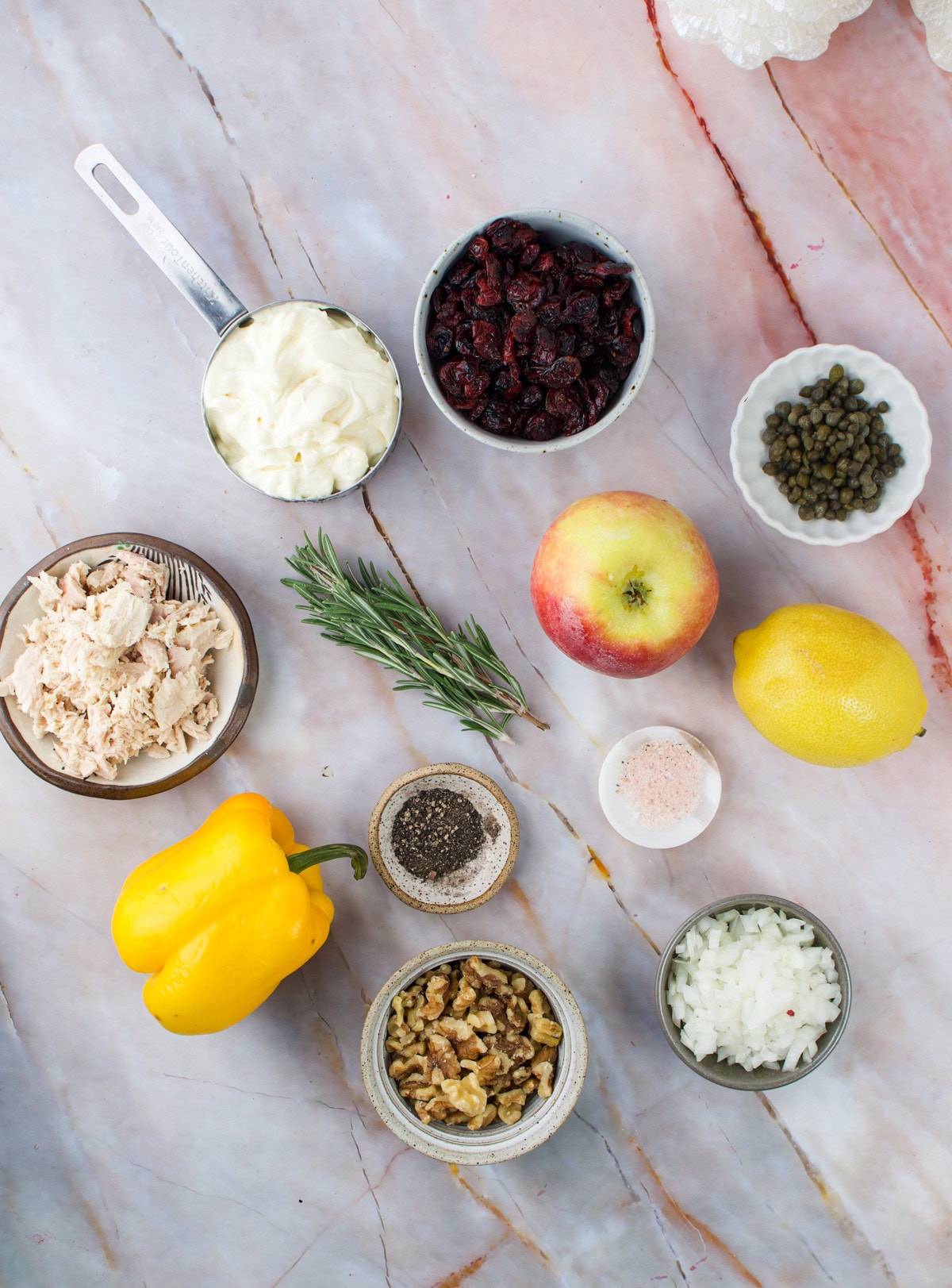 Various ingredients are arranged on a marble surface, including a yellow bell pepper, apple, lemon, fresh rosemary, mayonnaise, capers, dried cranberries, tuna, chopped onion, walnuts, and small bowls of salt and pepper.