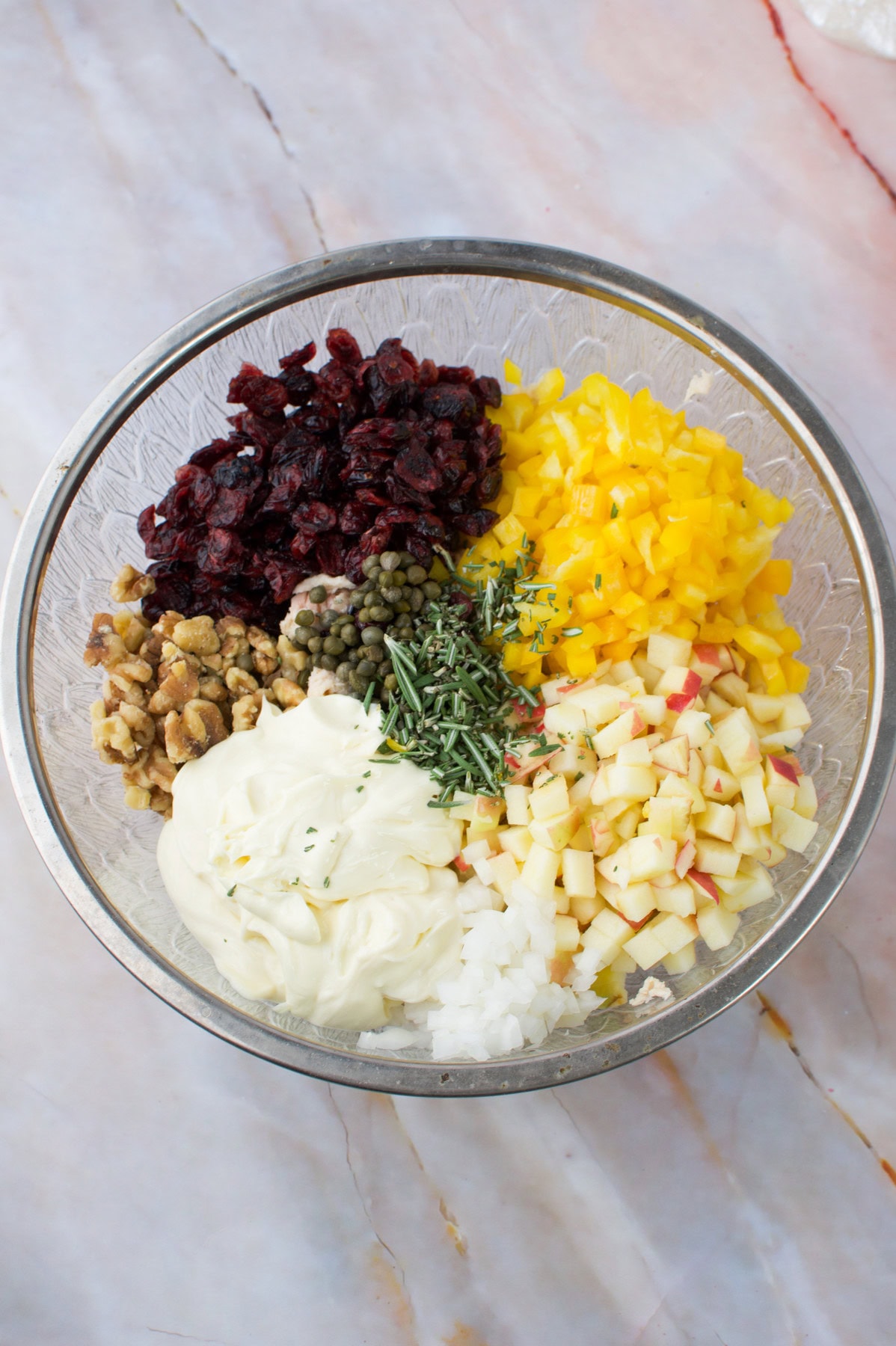 A glass bowl containing separate piles of dried cranberries, chopped yellow bell pepper, diced apples, walnuts, capers, chopped onion, fresh herbs, and a dollop of mayonnaise on a marble surface.