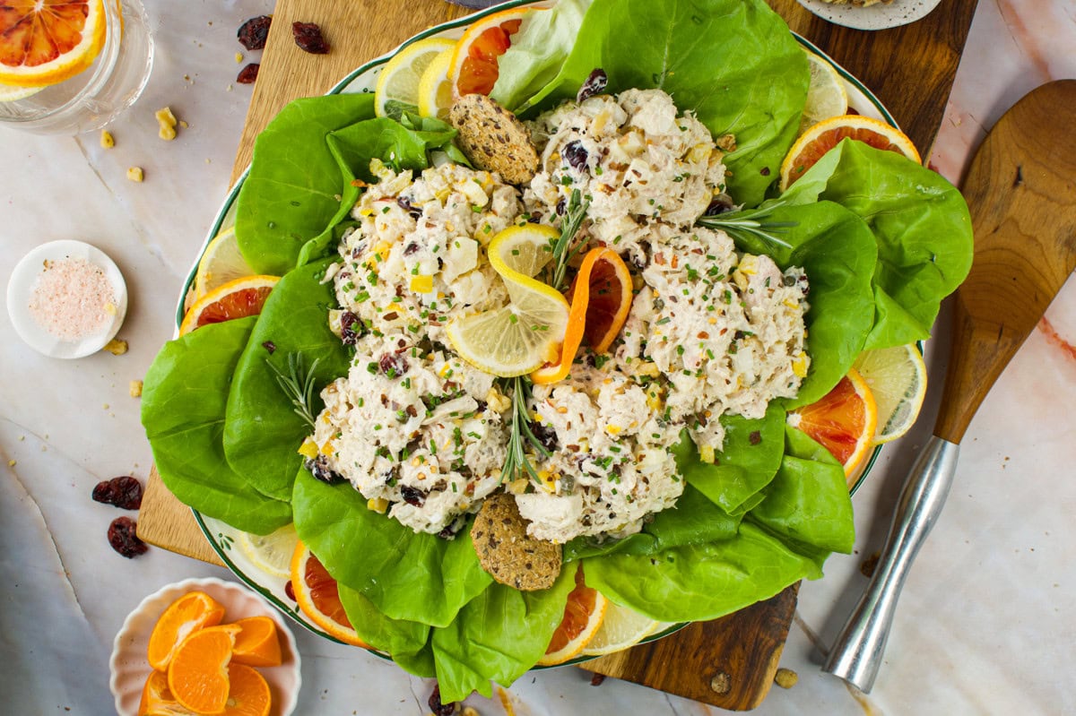 A plate of chicken salad with herbs and nuts, served on a bed of fresh green lettuce, garnished with lemon and orange slices, and accompanied by crackers. Wooden serving utensils and citrus fruits are nearby.
