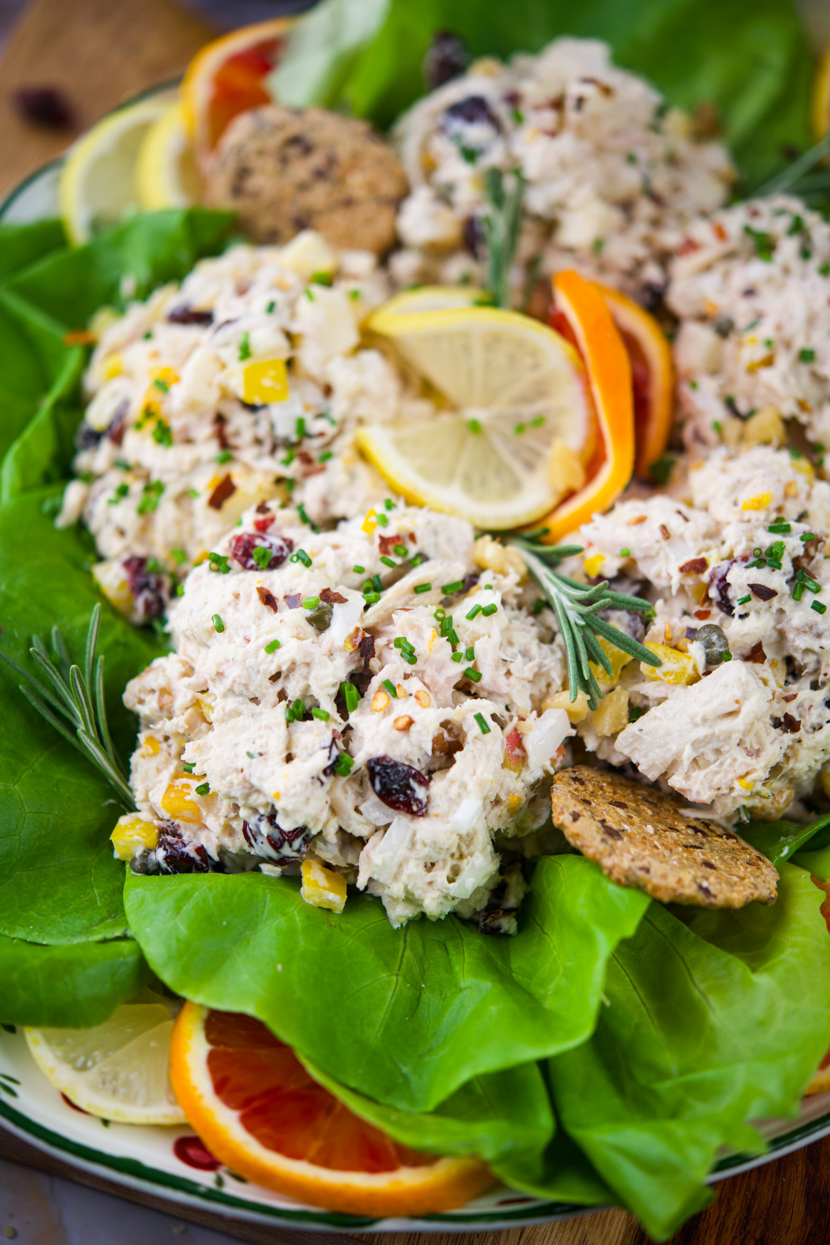 Cranberry tuna salad served on a bed of leafy greens, garnished with lemon and orange slices, fresh herbs, and crackers. The salad contains chunks of chicken, diced vegetables, and dried cranberries.