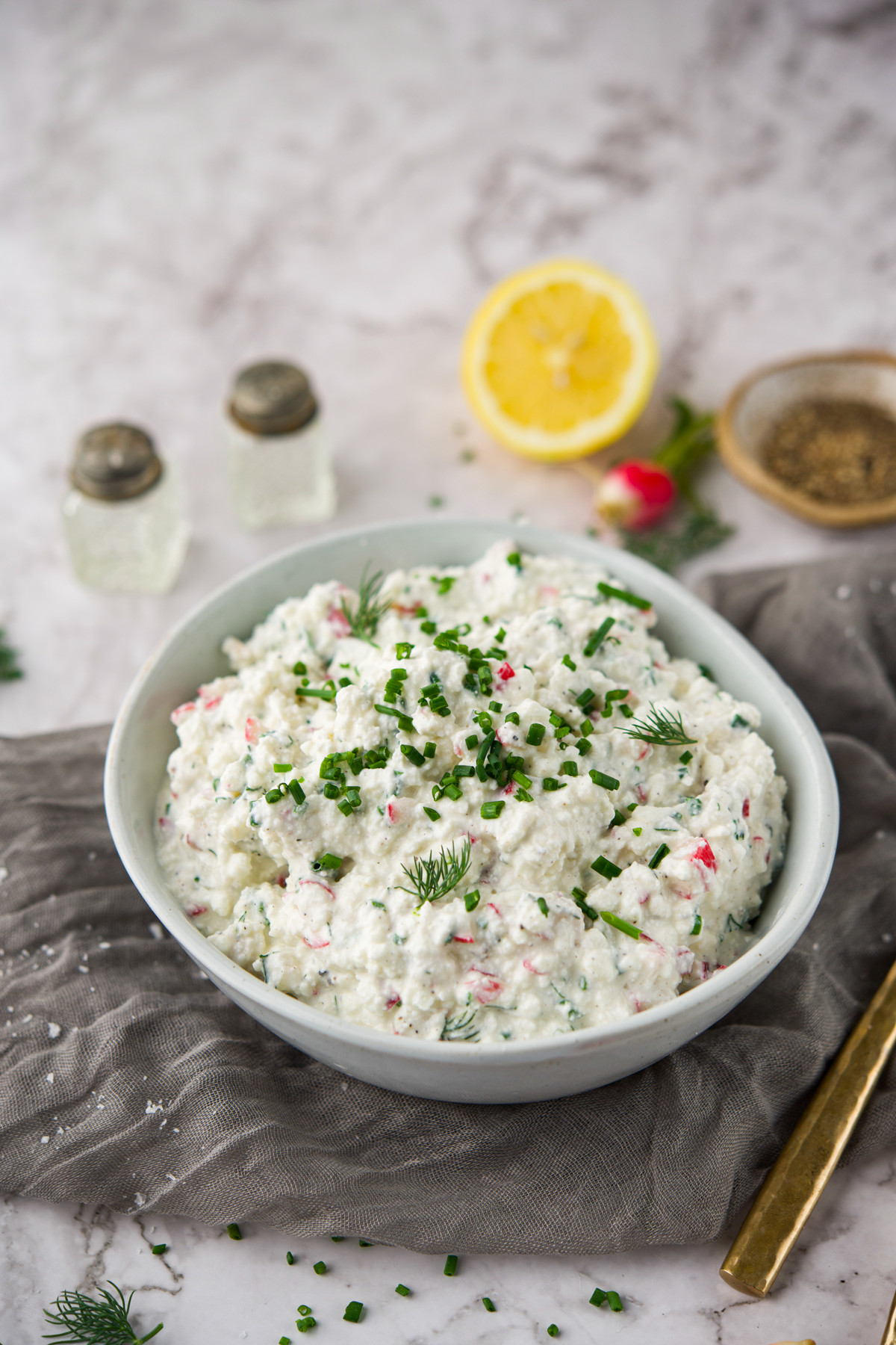 A bowl of creamy dip garnished with chopped chives and dill sits on a gray cloth, with a lemon half, salt and pepper shakers, and a gold spoon nearby on a marble surface.
