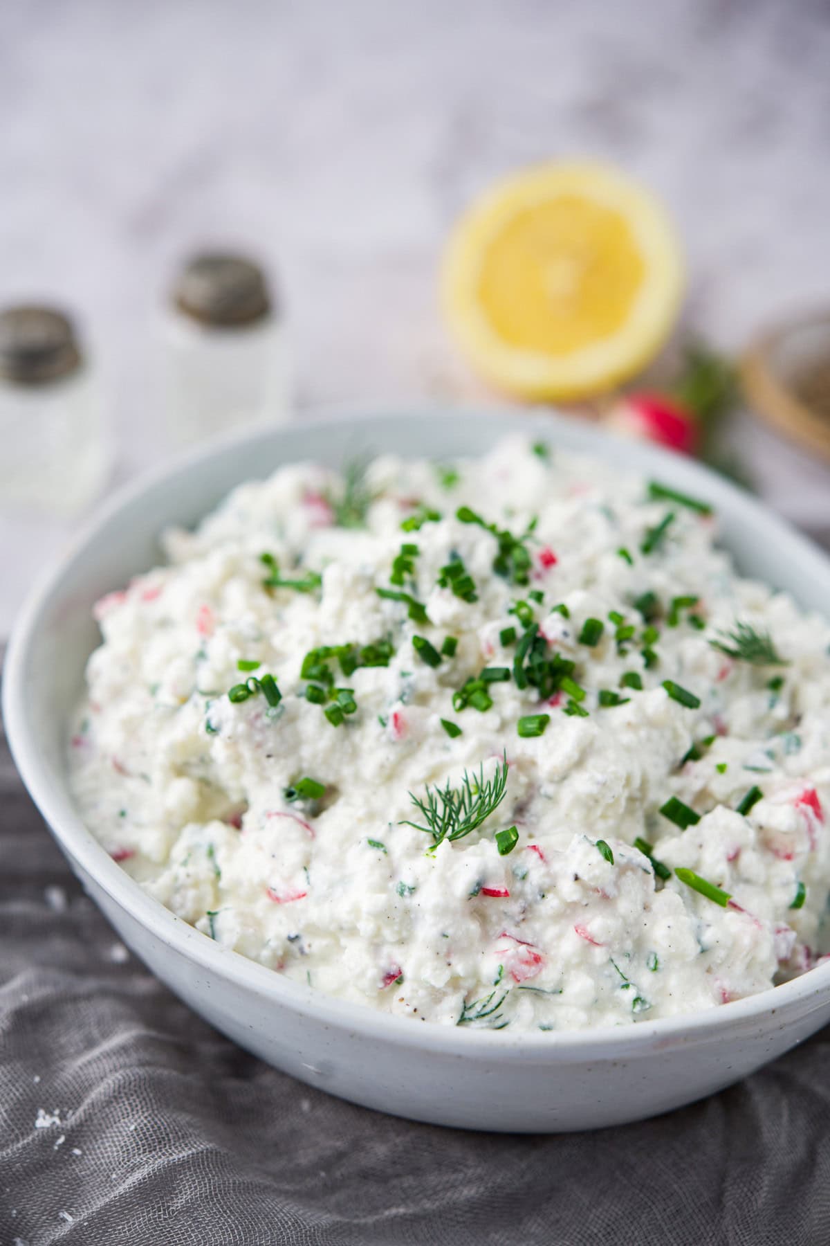 A white bowl filled with creamy Polish Gzik cottage cheese dip, garnished with chopped green chives and fresh dill. In the background, a halved lemon and two salt and pepper shakers are visible.