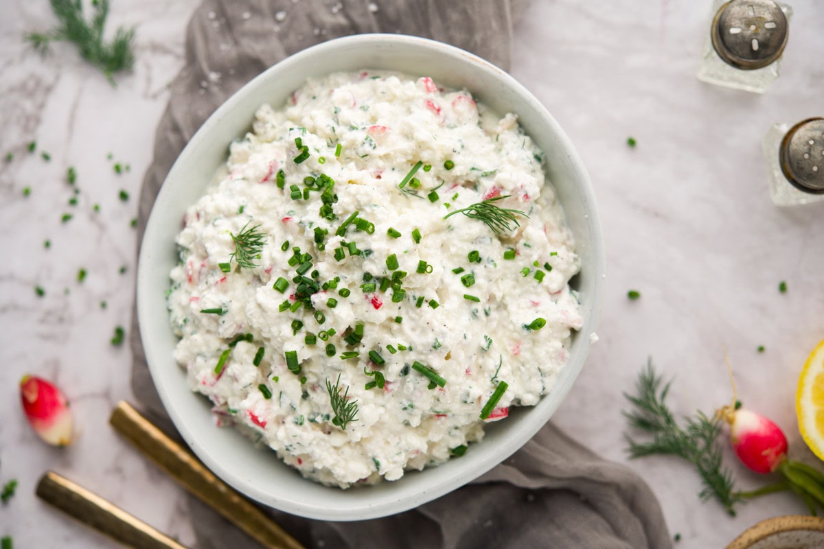A bowl of creamy dip garnished with chopped chives and dill sits on a marble surface, surrounded by gold utensils, radish slices, salt and pepper shakers, and fresh herbs.