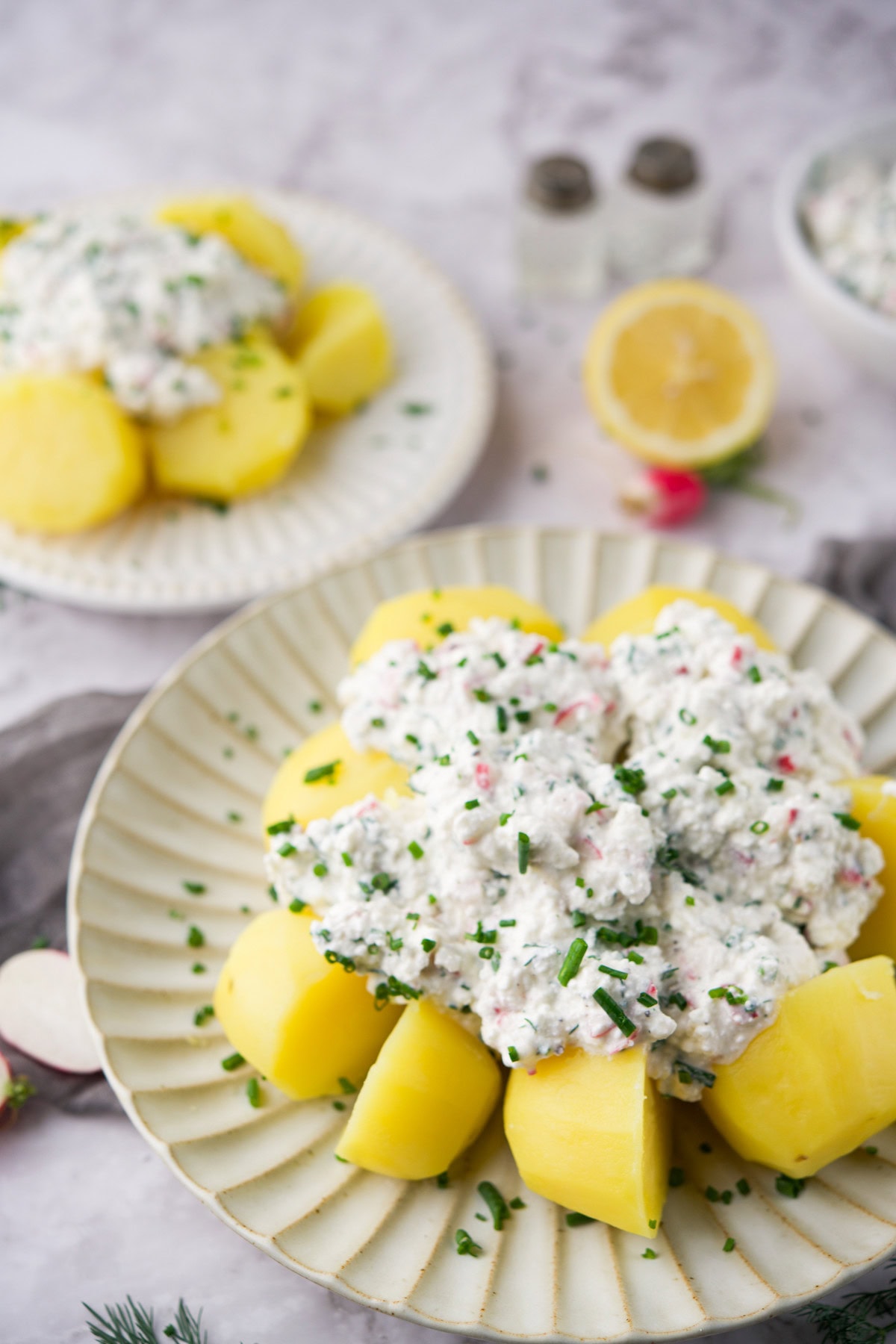 Plates of boiled potatoes topped with a creamy white salad garnished with chopped chives, with a halved lemon and salt and pepper shakers in the background on a light surface.