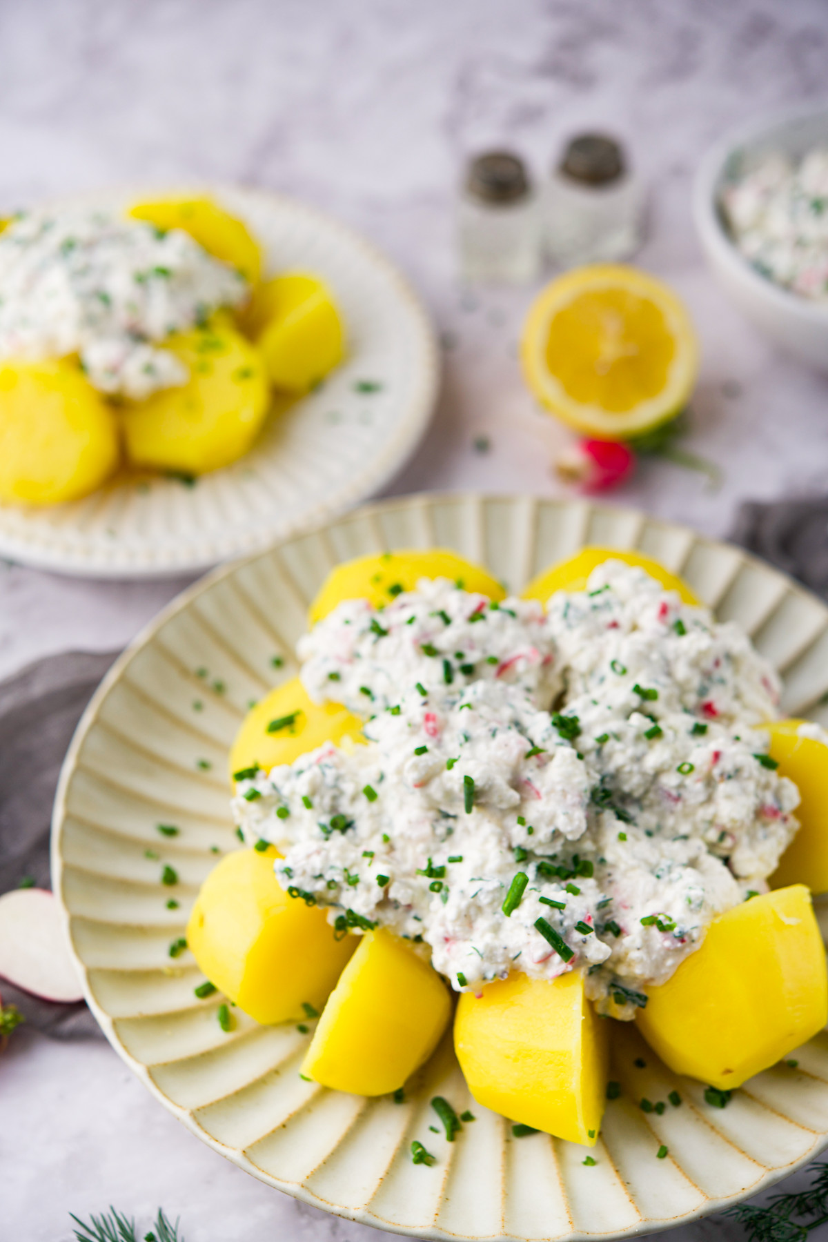 A plate of Pyry z Gzikiem (Potatoes with Farmer's Cheese Dip) garnished with chopped chives. A similar dish and ingredients are blurred in the background.