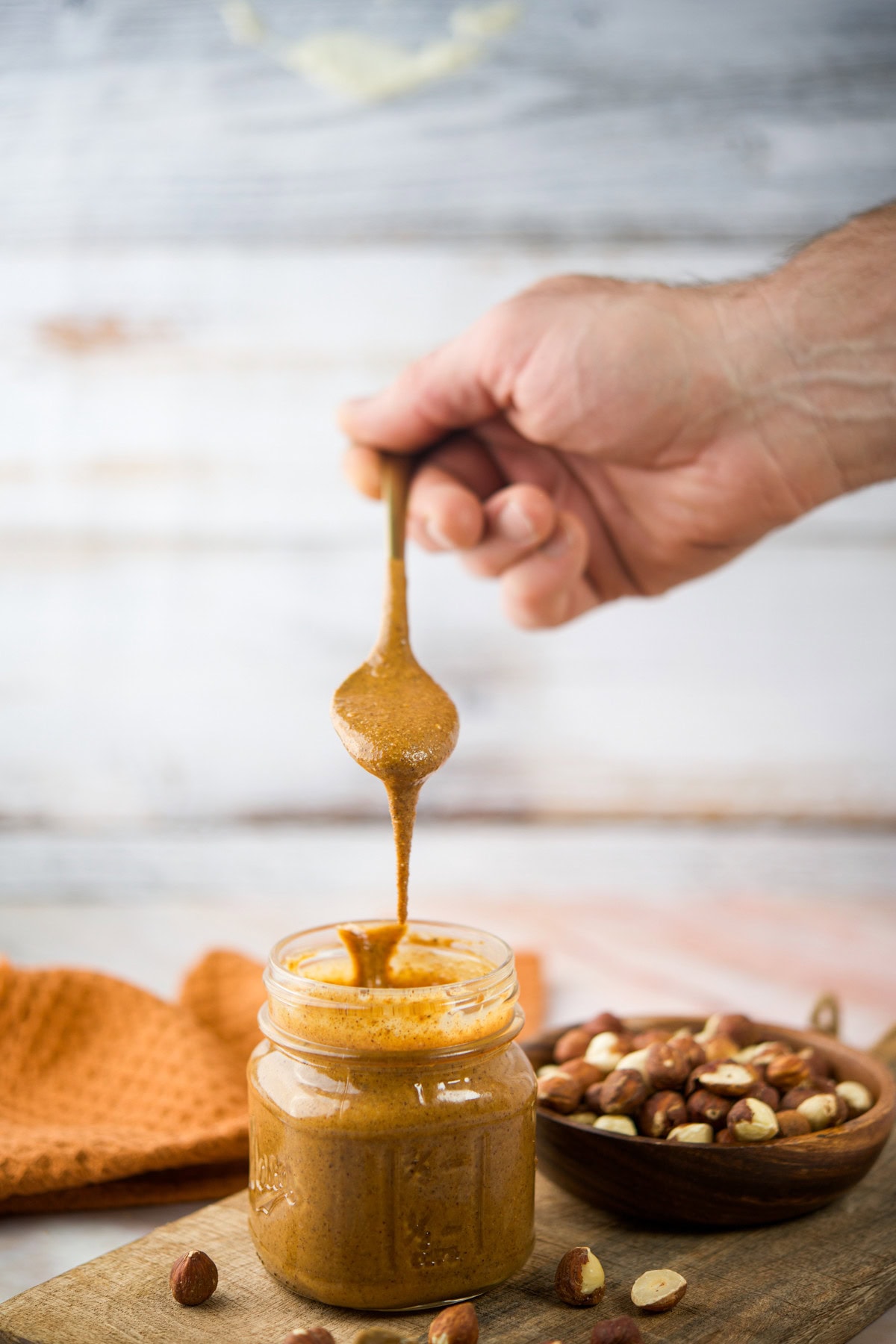 A hand holds a spoon dripping creamy nut butter into a glass jar, with shelled nuts scattered on a wooden board and a bowl of nuts in the background.