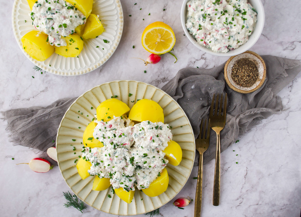 A plate of boiled yellow potatoes topped with a creamy salad garnished with chopped chives, surrounded by a lemon half, radishes, pepper, and utensils on a marble surface.