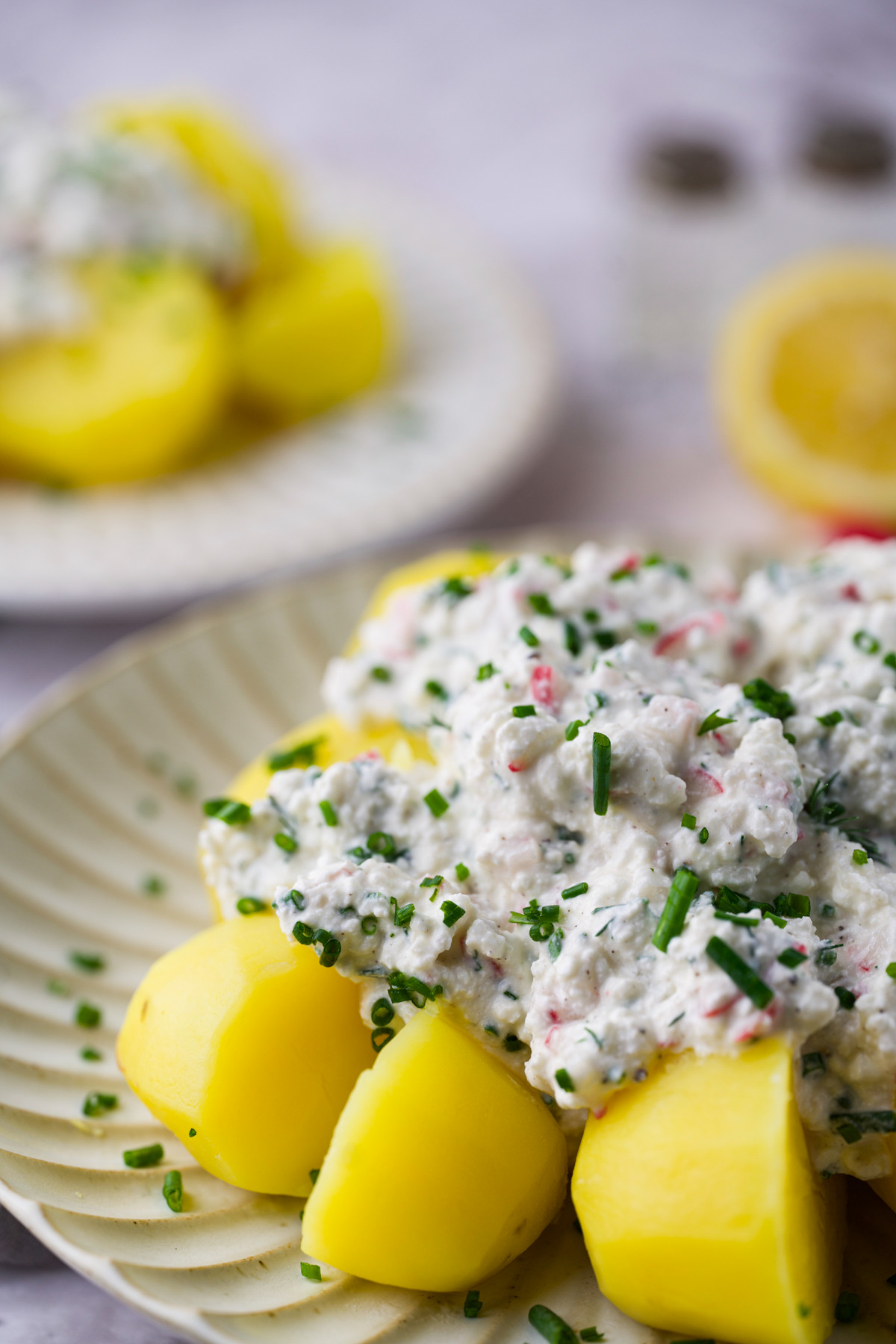 A plate of boiled yellow potatoes topped with a creamy salad mixture, garnished with chopped chives. A blurred plate and half a lemon are visible in the background.