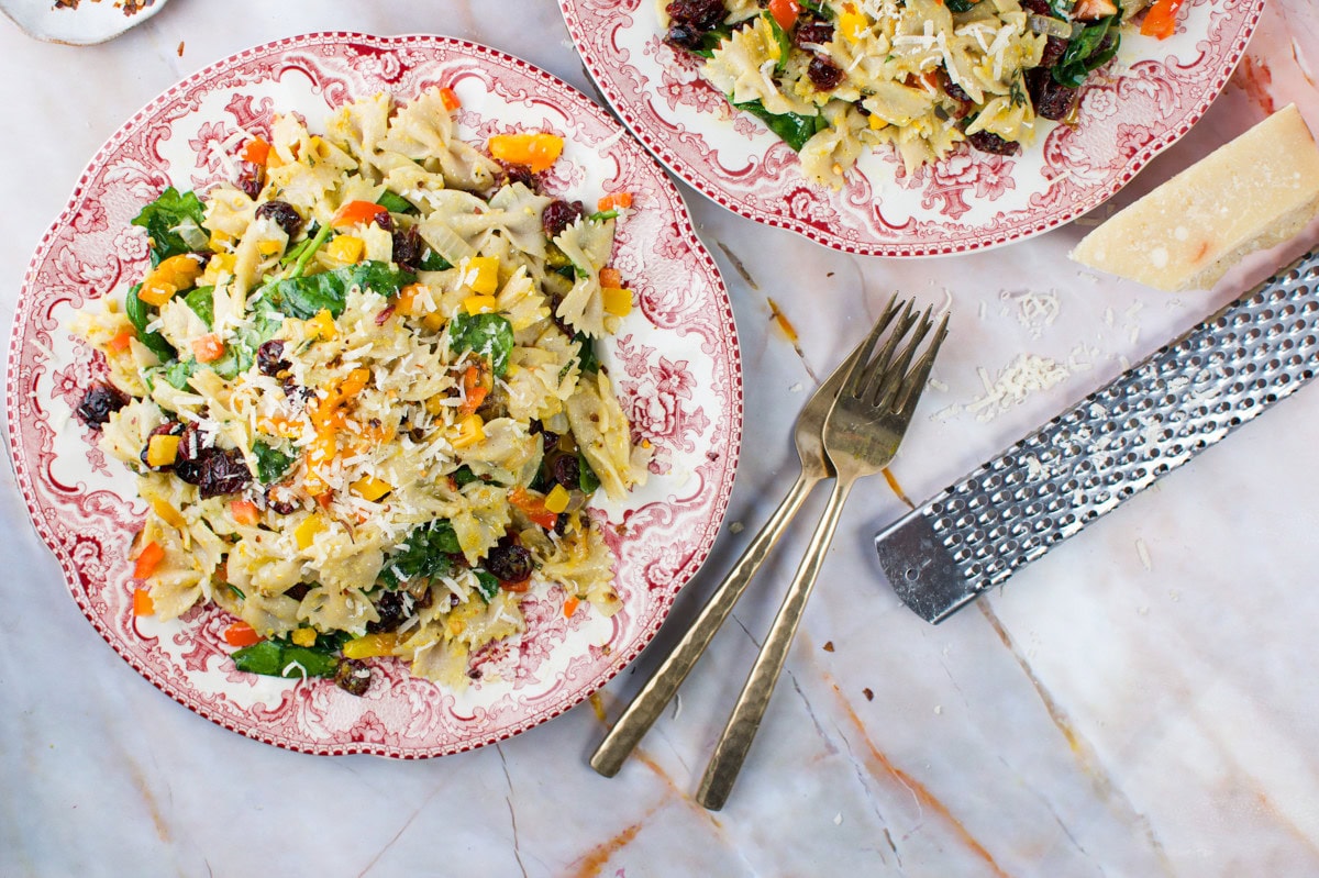 Two vintage plates filled with bowtie pasta salad featuring greens, yellow and red vegetables, and grated cheese. Two forks lie beside the plates, with a metal grater and a block of cheese nearby on a marble surface.