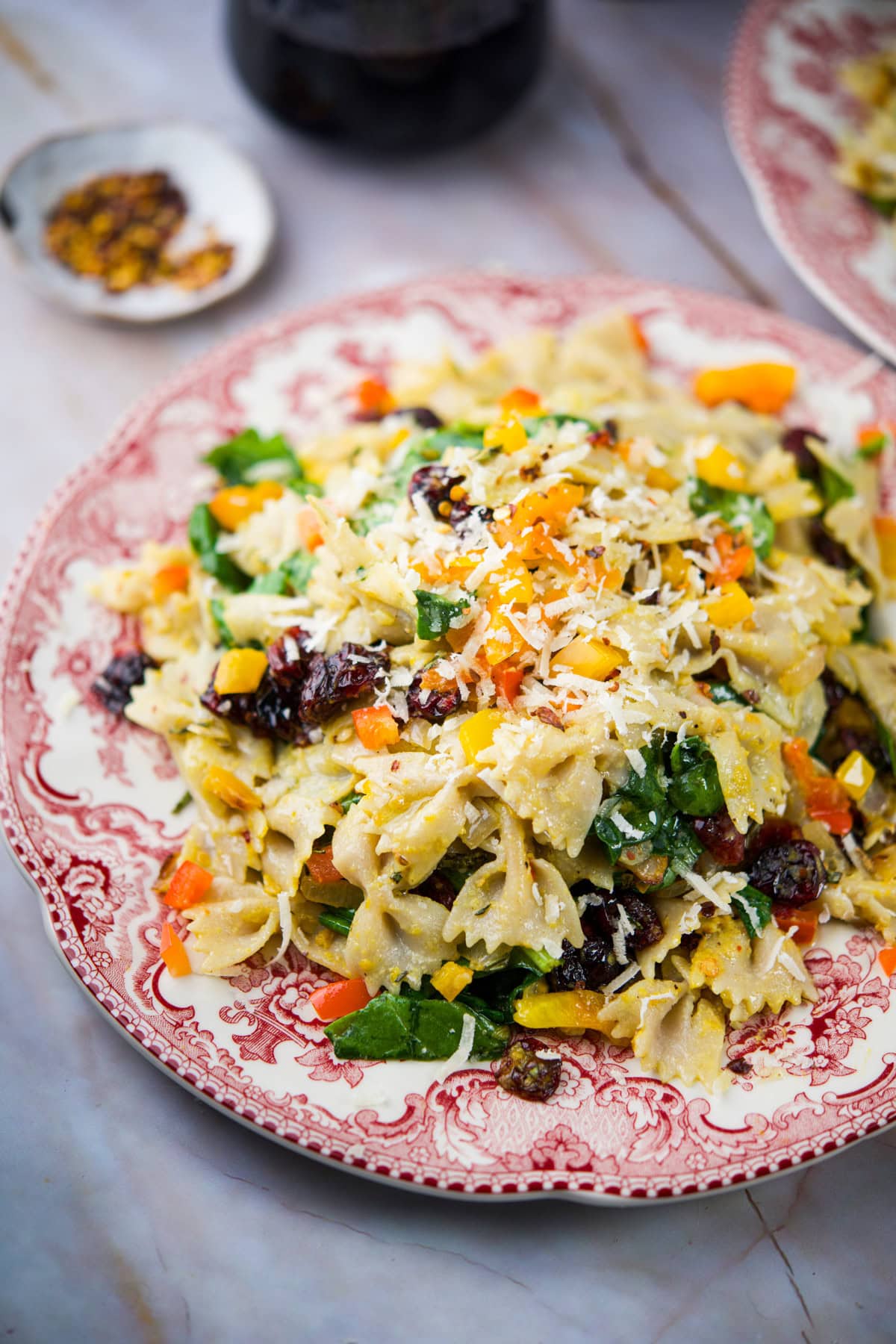 A plate of bowtie pasta mixed with spinach, dried cranberries, diced yellow and orange peppers, and topped with grated cheese, served on a patterned red and white plate.