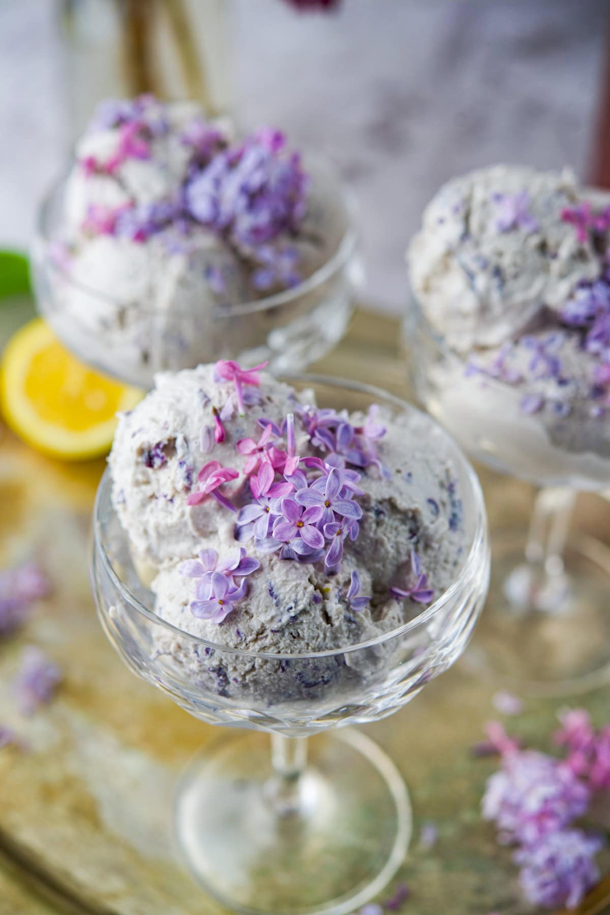 Scoops of creamy lilac ice cream served in elegant glass dessert cups, garnished with fresh purple and pink lilac blossoms