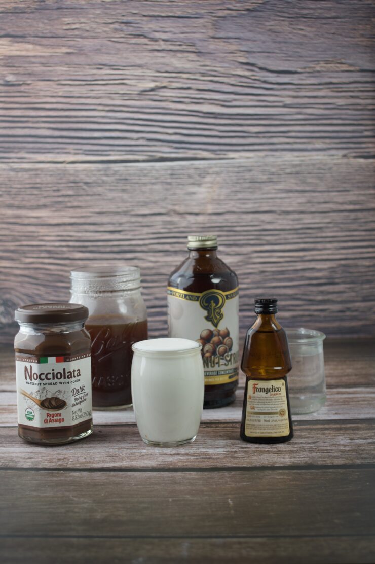 A glass of white liquid, jars of chocolate spread, an open jar with brown spread, a bottle of hazelnut syrup, a small bottle of liqueur, and an empty glass sit on a wooden surface with a wood-grain backdrop.