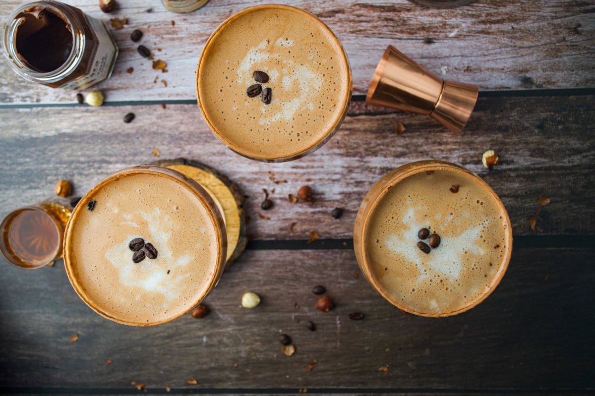 Three cups of frothy coffee topped with coffee beans are arranged on a wooden surface, surrounded by scattered coffee beans, a small jar, and a copper jigger.