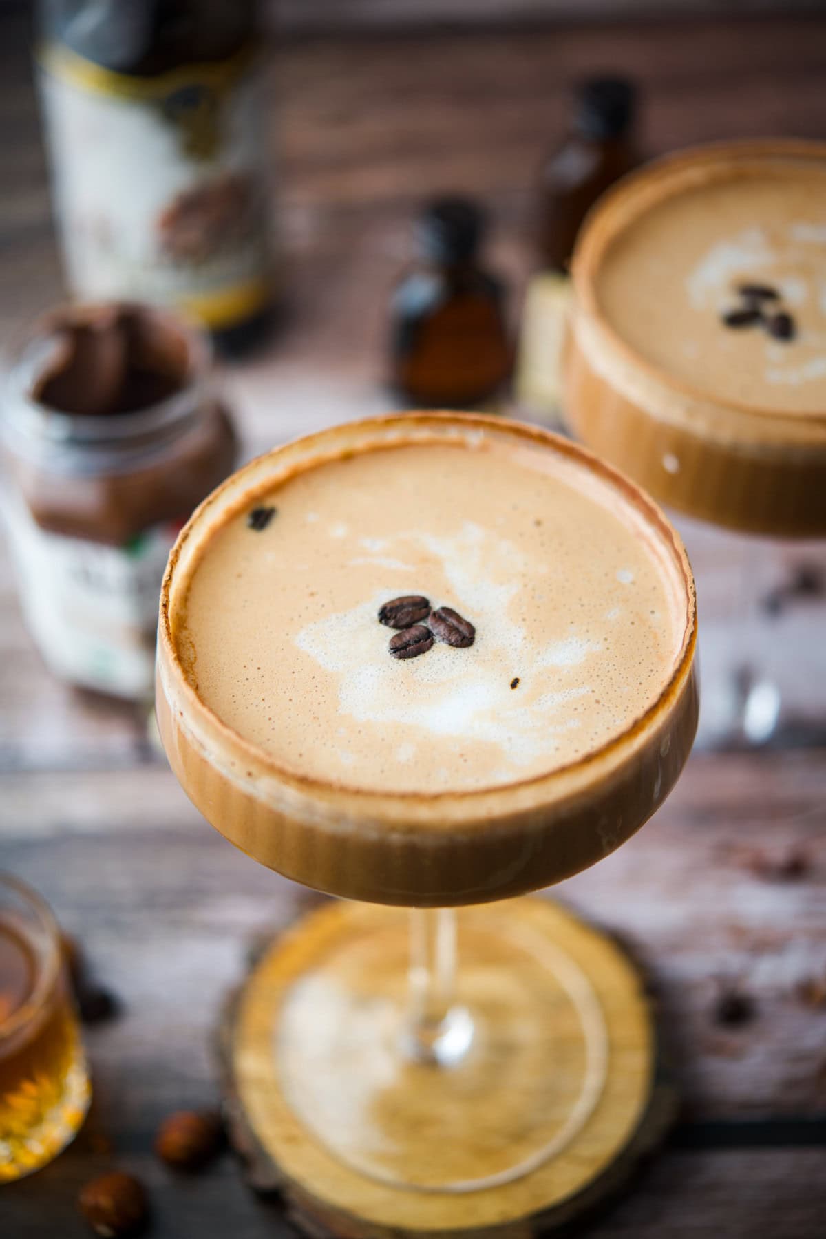 A close-up of a frothy espresso martini in a cocktail glass, garnished with three coffee beans. The drink sits on a wooden coaster, with blurred bottles and jars in the rustic background.