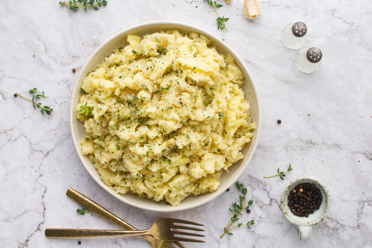 A bowl of mashed potatoes garnished with herbs sits on a marble surface, surrounded by a gold fork, thyme sprigs, black pepper, garlic, and salt and pepper shakers.