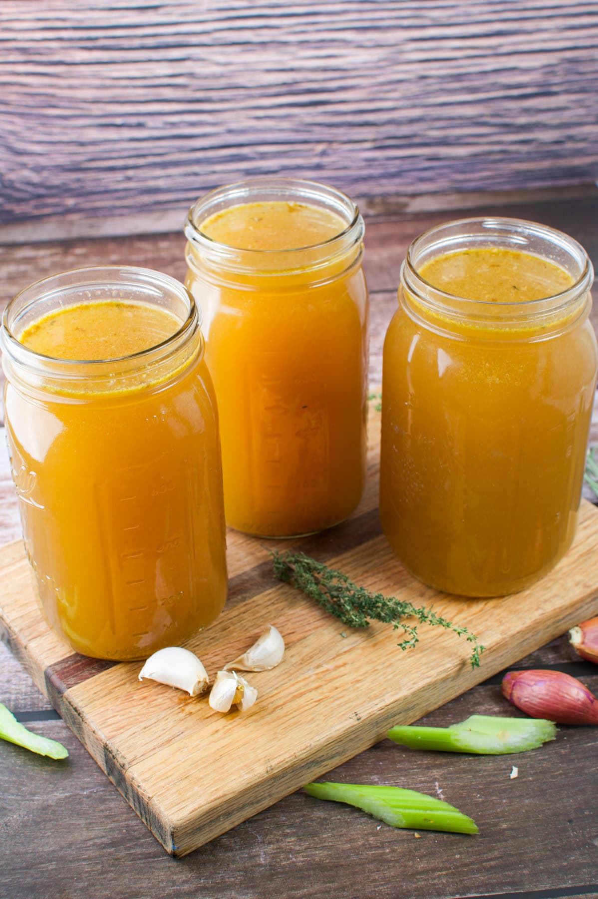 Three mason jars filled with golden-brown broth sit on a wooden board, surrounded by garlic cloves, fresh thyme, green onions, and shallots on a rustic wooden table.