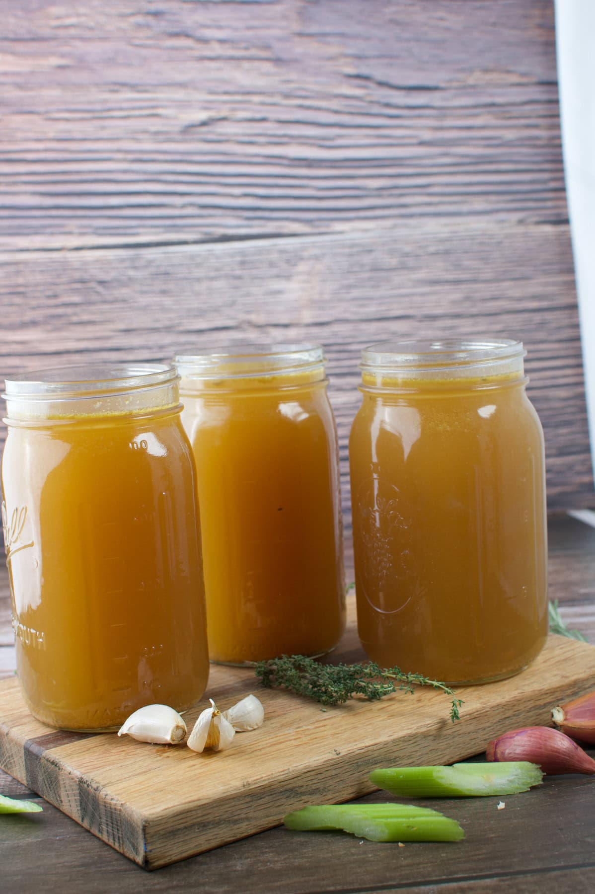 Three mason jars filled with brown broth sit on a wooden board, surrounded by garlic cloves, fresh thyme, celery, and shallots, with a wooden background behind them.