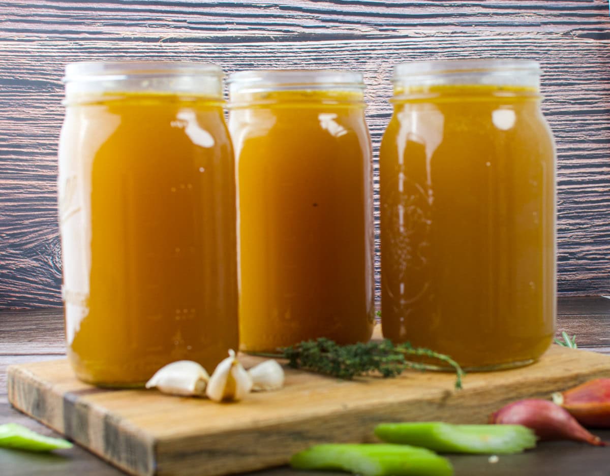 Three large mason jars filled with golden brown broth sit on a wooden board, surrounded by garlic cloves, fresh thyme, celery, and shallots, with a rustic wooden background behind them.