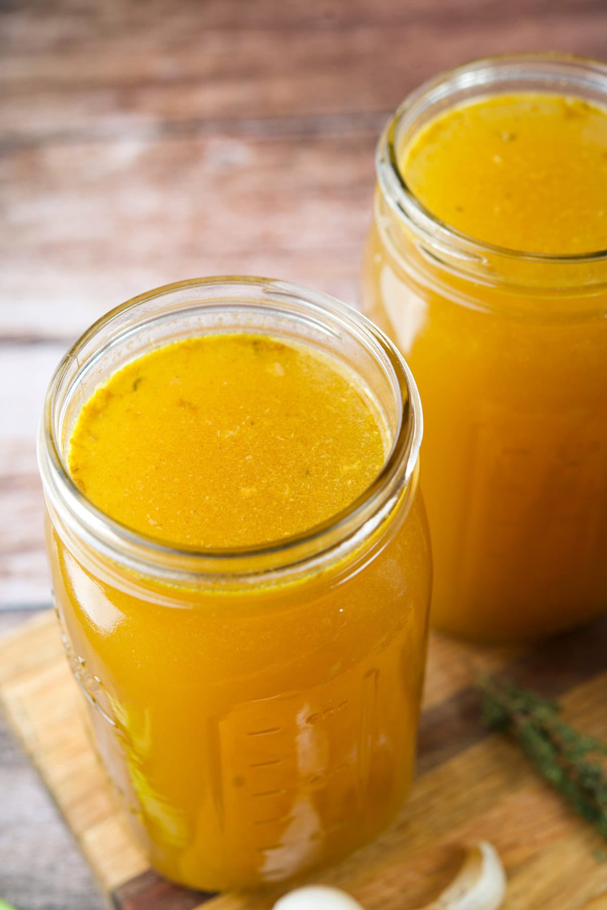 Two glass jars filled with Leftover Turkey Bone Broth sit on a wooden surface, with a clove of garlic and a sprig of herbs nearby.
