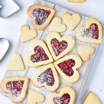 A wooden board displays heart-shaped Gluten-Free Linzer Cookies some with jam centers and powdered sugar, surrounded by plain heart cookies and heart-shaped cookie cutters on a white surface.