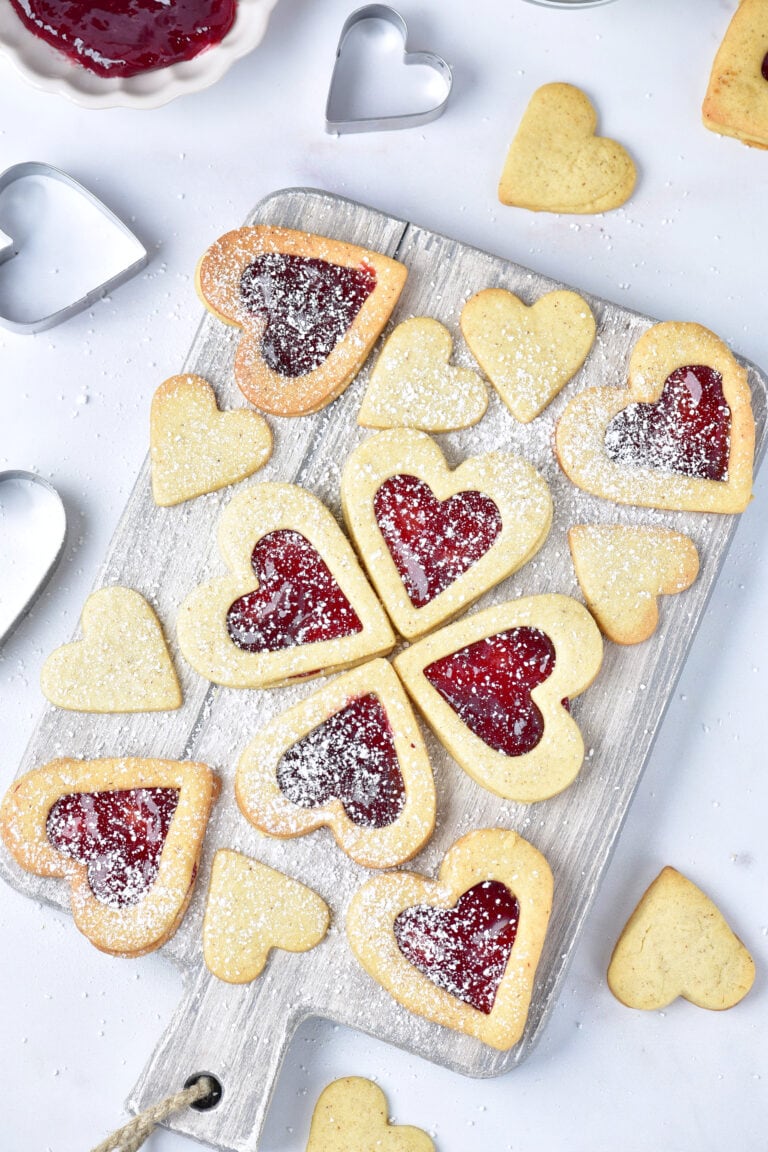 A wooden board displays heart-shaped Gluten-Free Linzer Cookies some with jam centers and powdered sugar, surrounded by plain heart cookies and heart-shaped cookie cutters on a white surface.