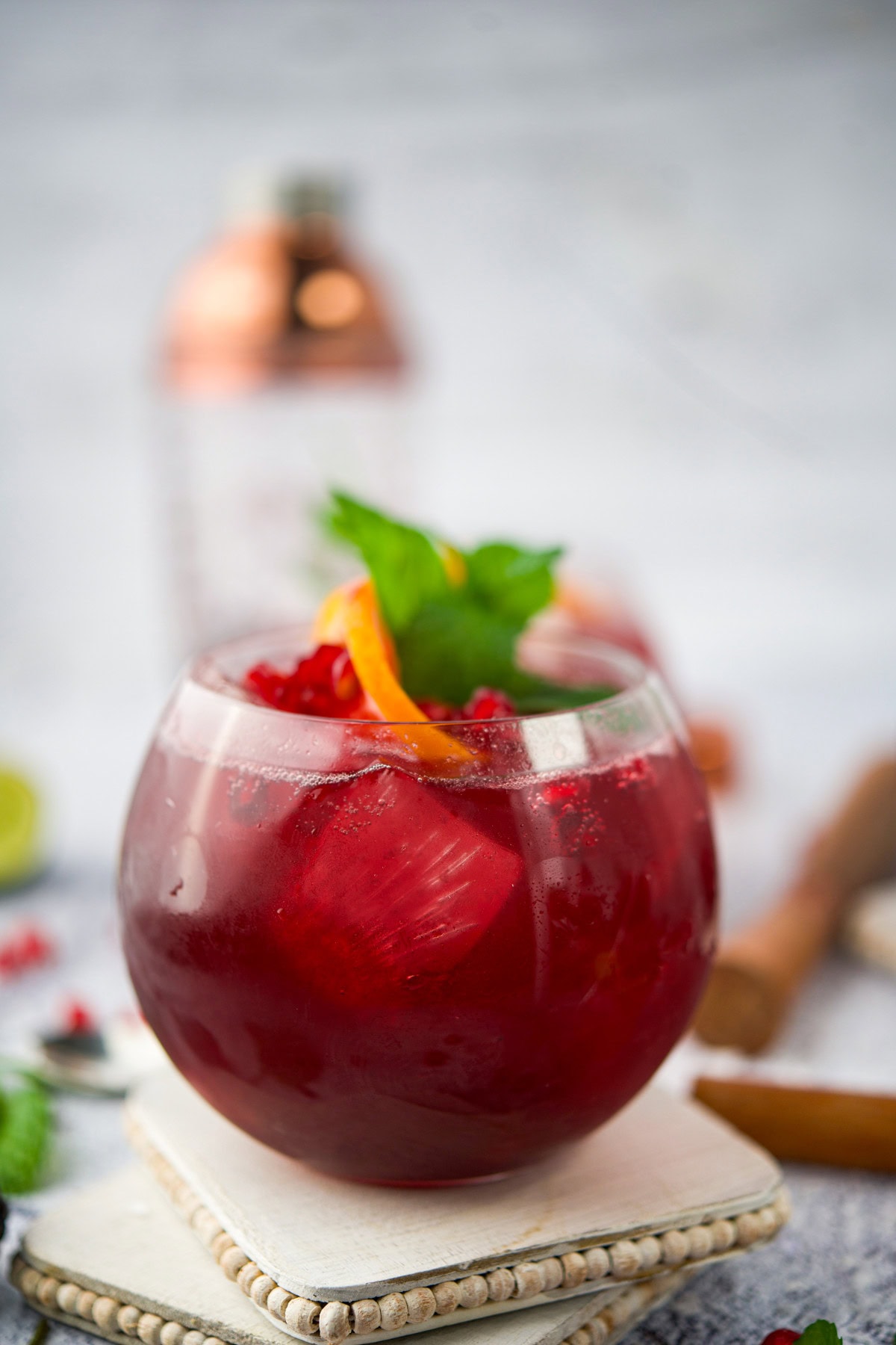 A round glass filled with a Pomegranate Blackberry Cocktail, garnished with an orange peel twist and fresh mint leaves, sits on a white coaster with a cocktail shaker blurred in the background.