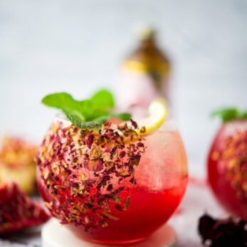 A round glass of vibrant Rose Pomegranate Mocktail Spritz garnished with rose petals, a lemon slice, and fresh mint, sits on a white coaster. Colorful straws and pomegranate seeds are scattered nearby.
