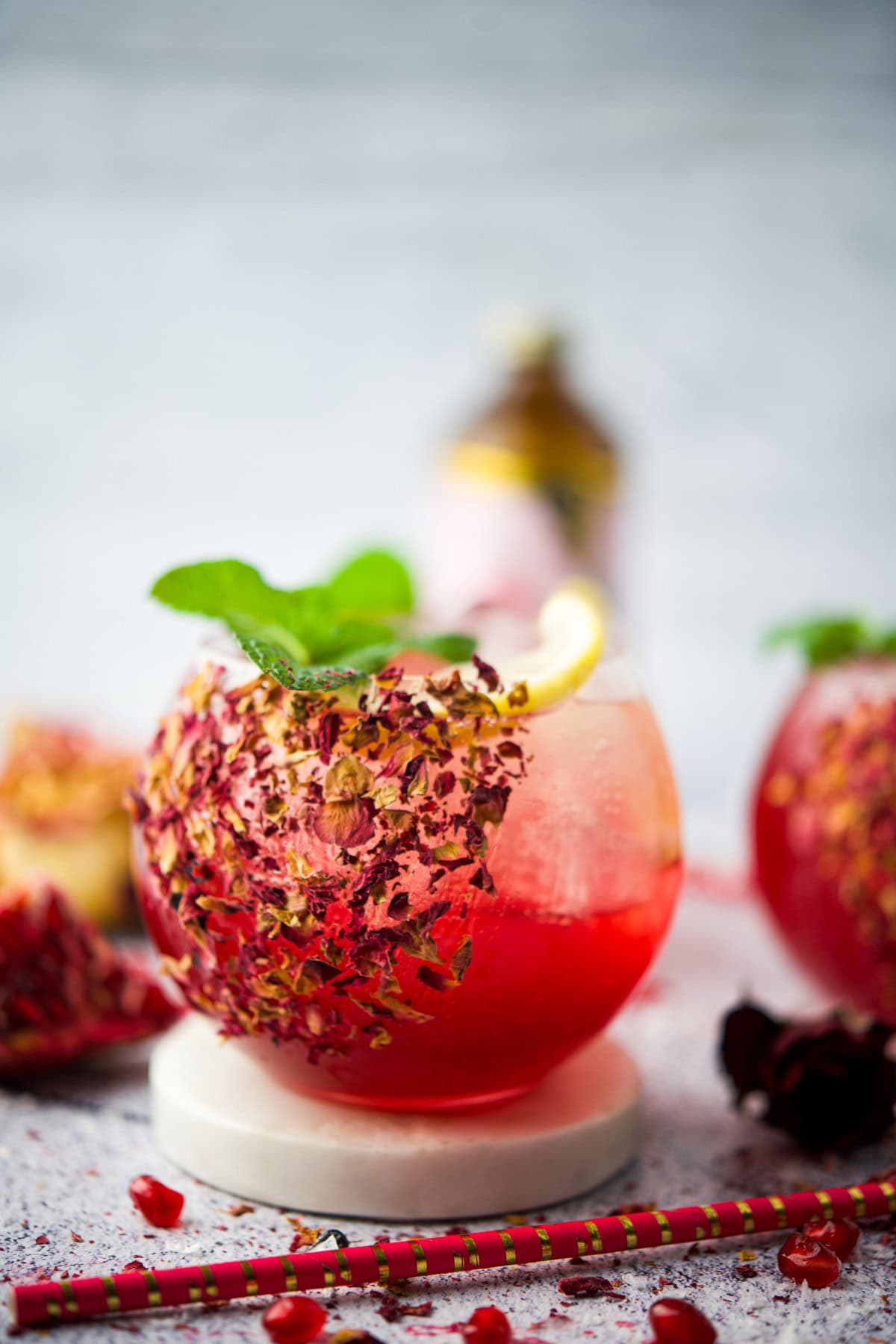 A round glass of vibrant Rose Pomegranate Mocktail Spritz garnished with rose petals, a lemon slice, and fresh mint, sits on a white coaster. Colorful straws and pomegranate seeds are scattered nearby.