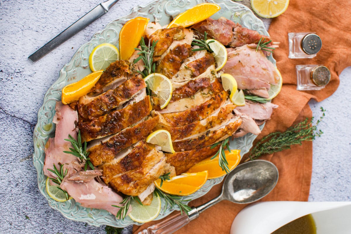 Sliced roasted turkey breast garnished with lemon and orange wedges, thyme, and rosemary on a decorative platter, with cutlery and salt and pepper shakers nearby on an orange napkin.