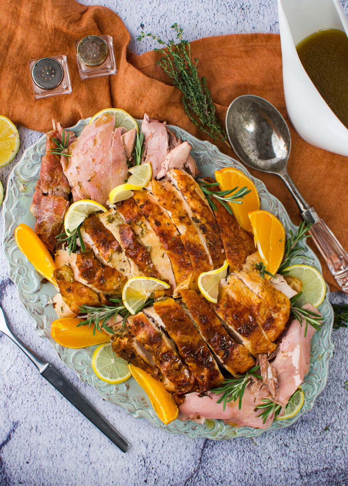 A platter of sliced roasted turkey garnished with lemon, orange wedges, and rosemary. A carving fork and knife, serving spoon, gravy boat, and salt and pepper shakers are nearby on a textured surface with an orange cloth.