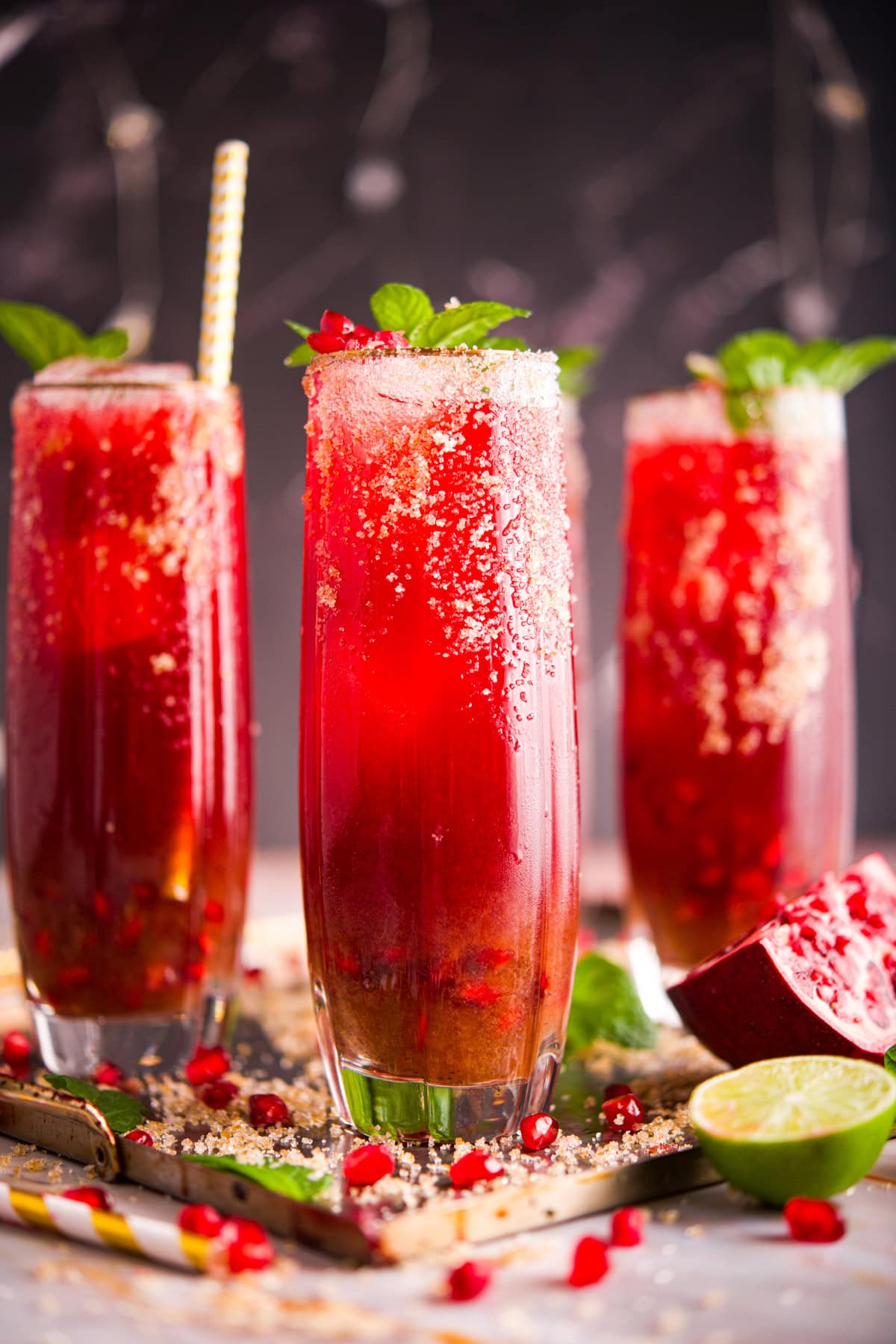 Three tall glasses of vibrant Pomegranate Mezcal Paloma cocktails, garnished with mint leaves and sugar rims, stand on a tray. Pomegranate seeds, lime halves, and a gold-striped straw add a festive touch to the scene.