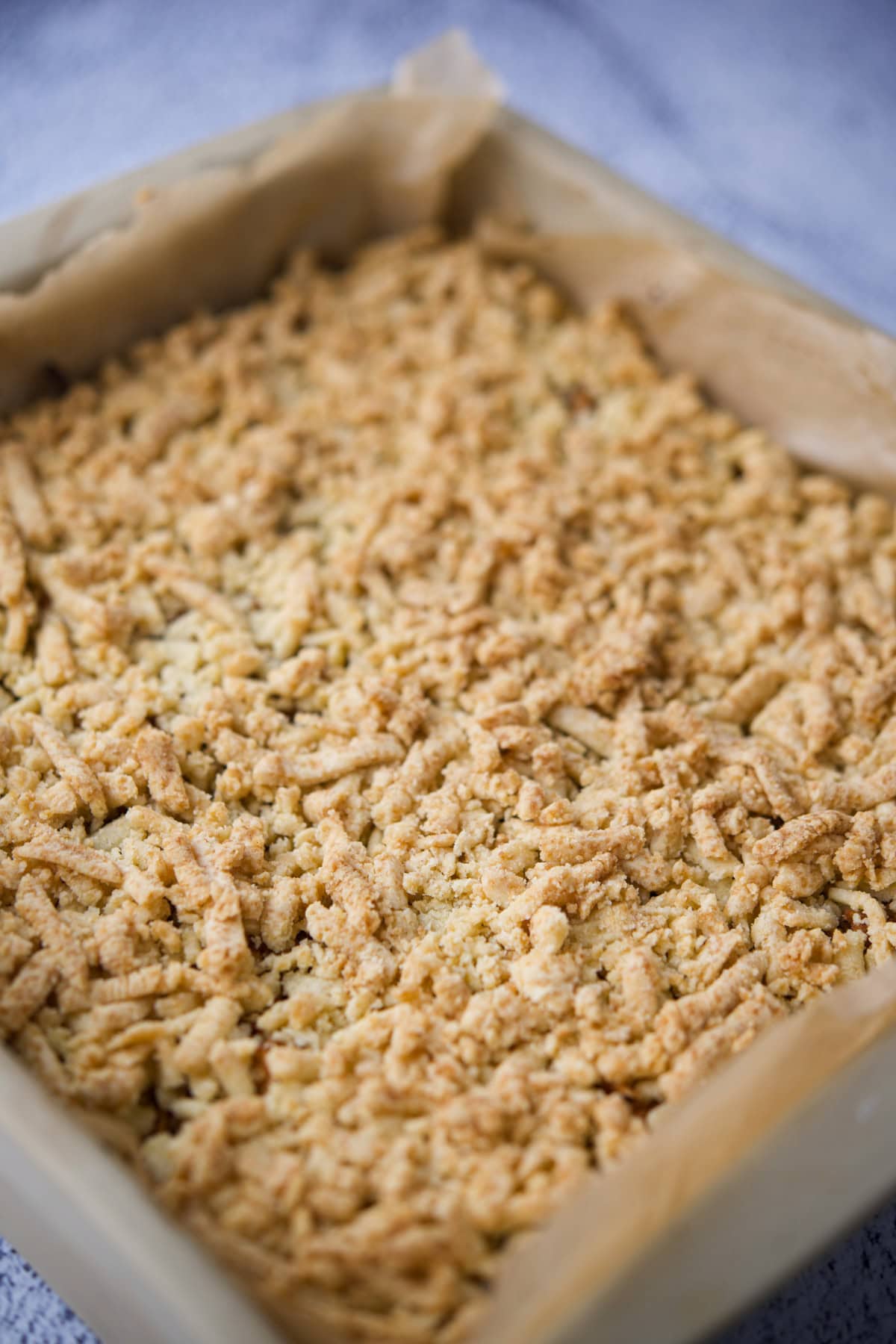 A close-up of a square baking pan lined with parchment paper and filled with freshly baked crumbly streusel or crumble topping, likely for a dessert such as bars or coffee cake.