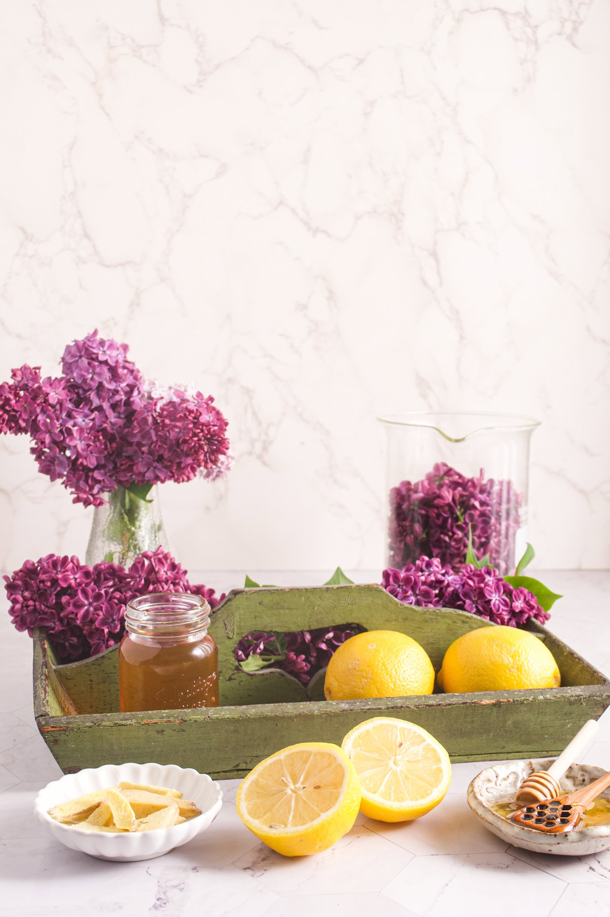 A still life scene with a green tray holding lemons and a jar of honey, surrounded by sliced lemons, a honey dipper, and purple lilacs in glass vases, set against a light marble background.