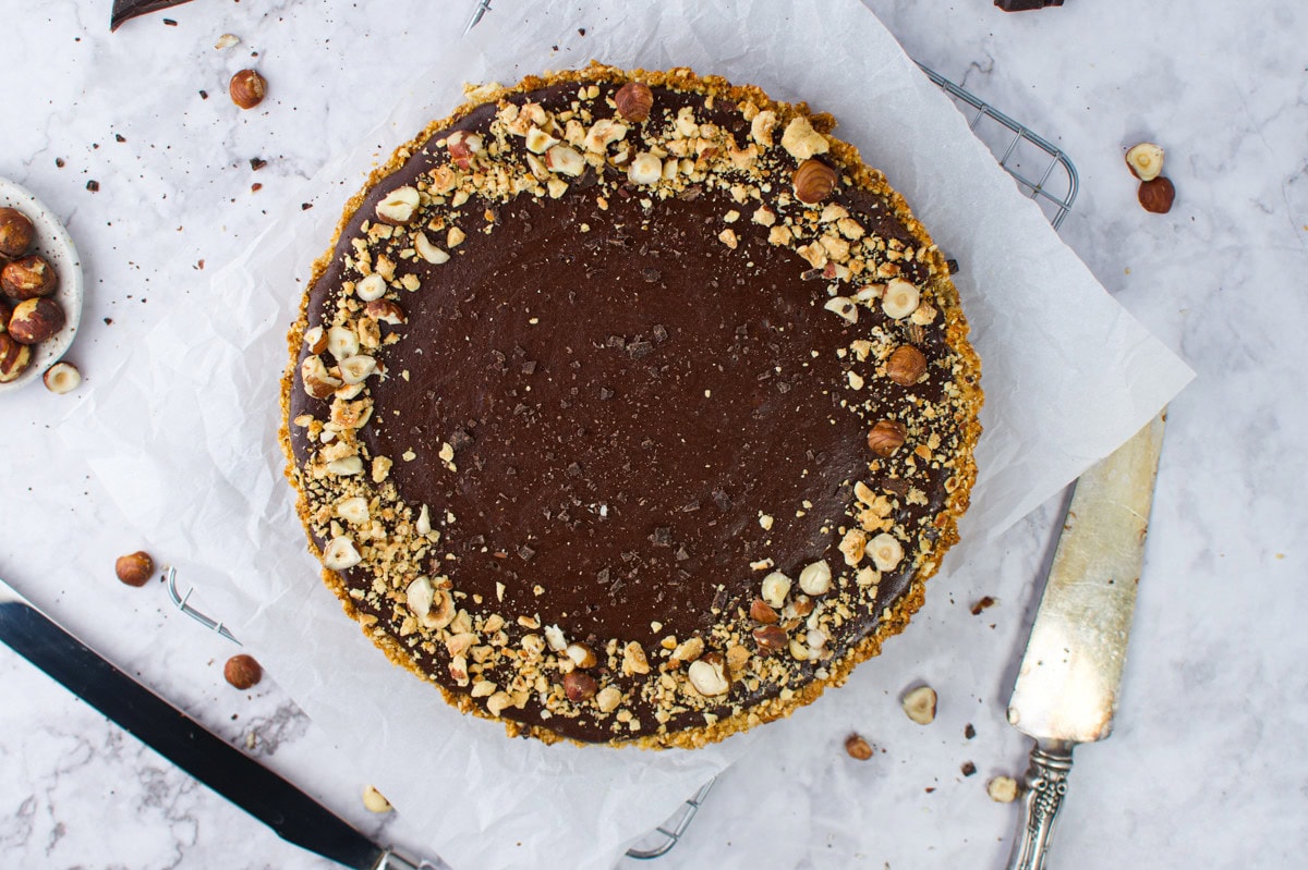 A round chocolate tart with a nutty crust, topped with chopped hazelnuts, sits on parchment paper. A knife and pie server are beside it, with scattered nuts on a light marble surface.