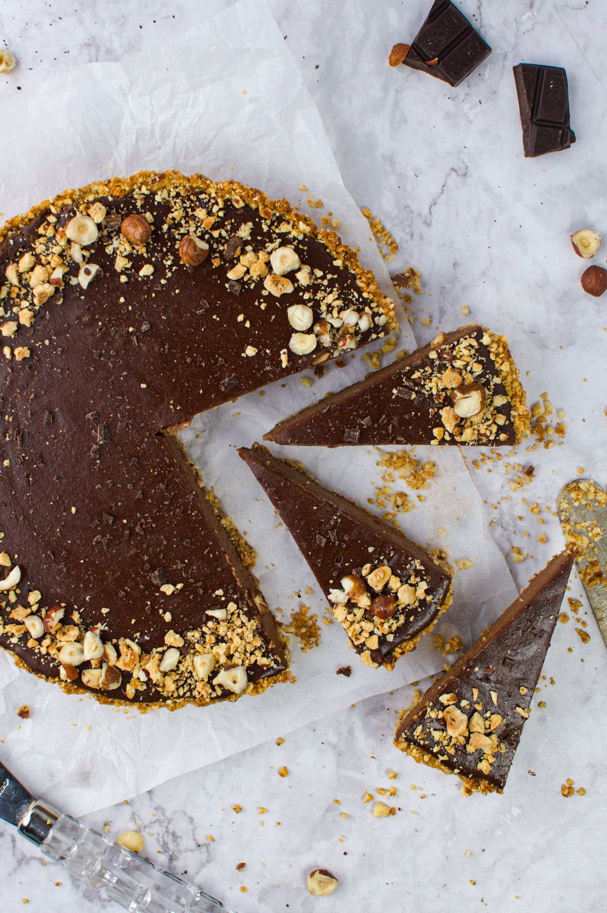 A chocolate tart with a nutty crust sits on parchment paper. Three slices have been cut and separated from the tart. Chopped hazelnuts are sprinkled on top, and chocolate pieces and crumbs are scattered nearby.