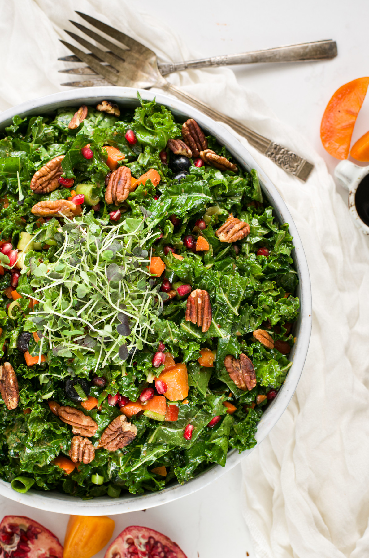 A bowl of kale salad topped with pecans, pomegranate seeds, shredded carrots, and microgreens, with a fork and orange slices nearby on a white tablecloth.