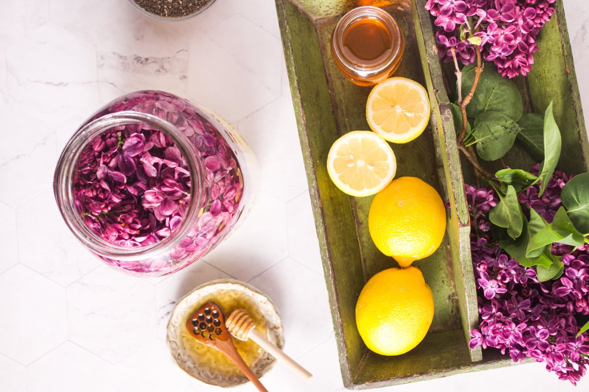 A jar of purple lilac flowers, a green tray with lemons, lemon slices, lilac flowers, a small jar of honey, and a bowl with honey and a honey dipper arranged on a white surface.