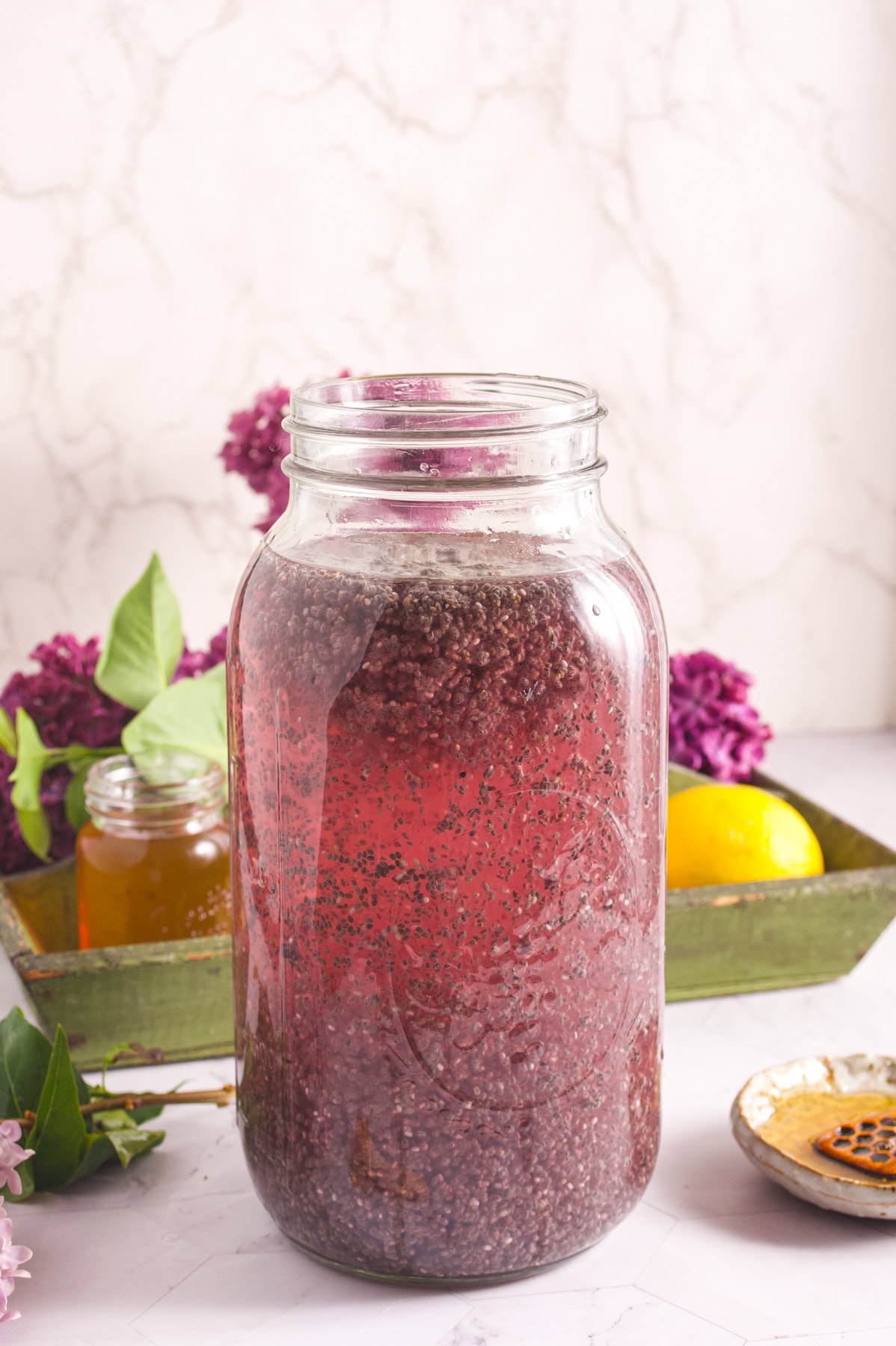 A large glass jar filled with a reddish-pink chia seed drink sits on a table. In the background are purple flowers, a lemon, a small jar of honey, and a green tray.
